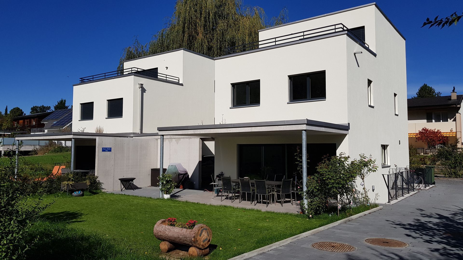 Modernes weißes Gebäude mit Flachdach, Terrasse und grünem Rasen an einem sonnigen Tag.