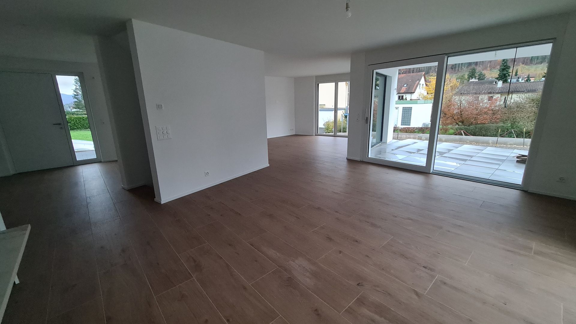 Leerer Raum mit Holzoptikboden, großen Fenstern und einer Tür, die auf eine Terrasse mit grünem Rasen führt.