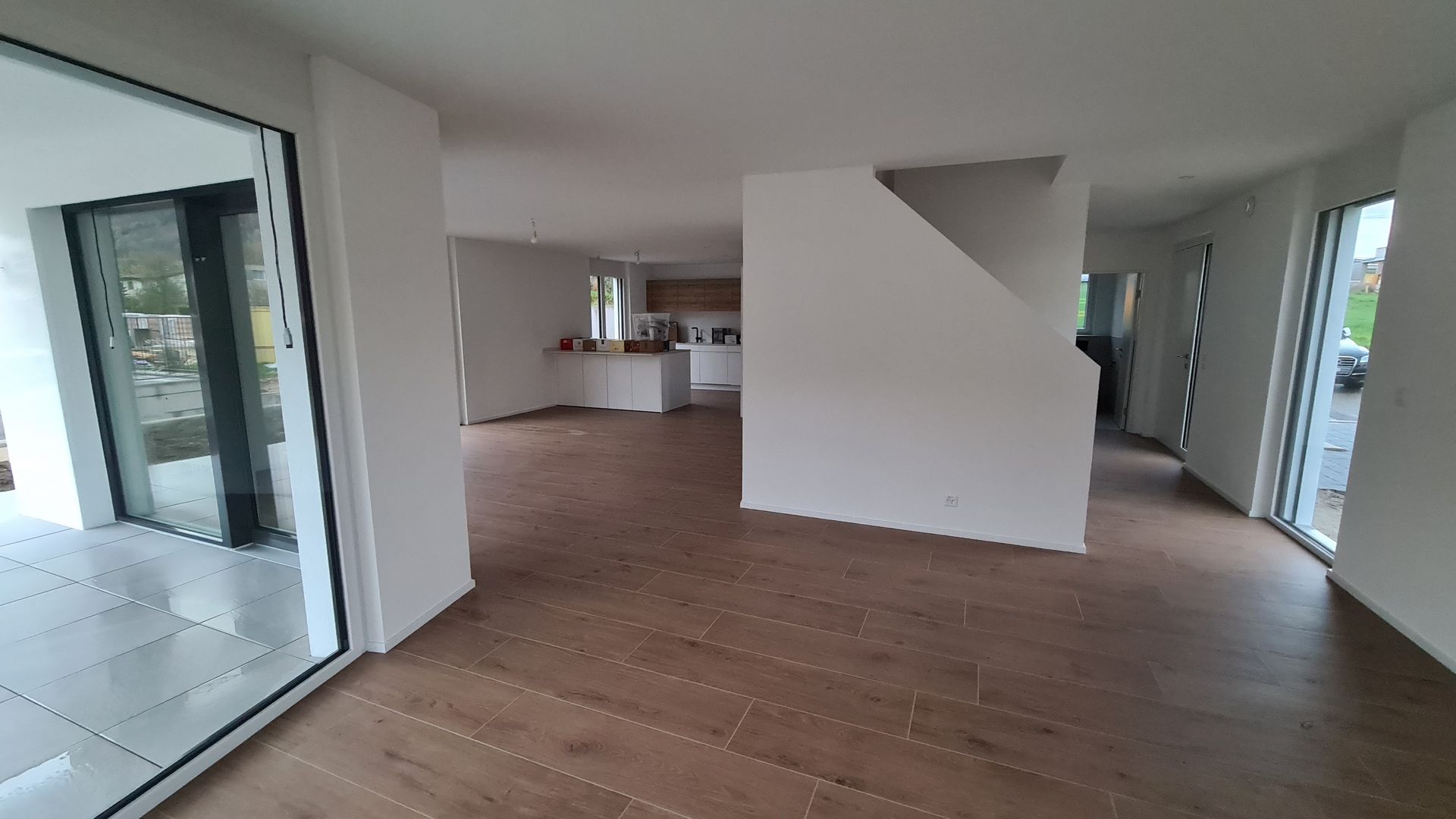 Geräumiges, helles Wohnzimmer mit Hartholzböden, weißen Wänden und großen Glastüren. Im Hintergrund ist eine Küche zu sehen.