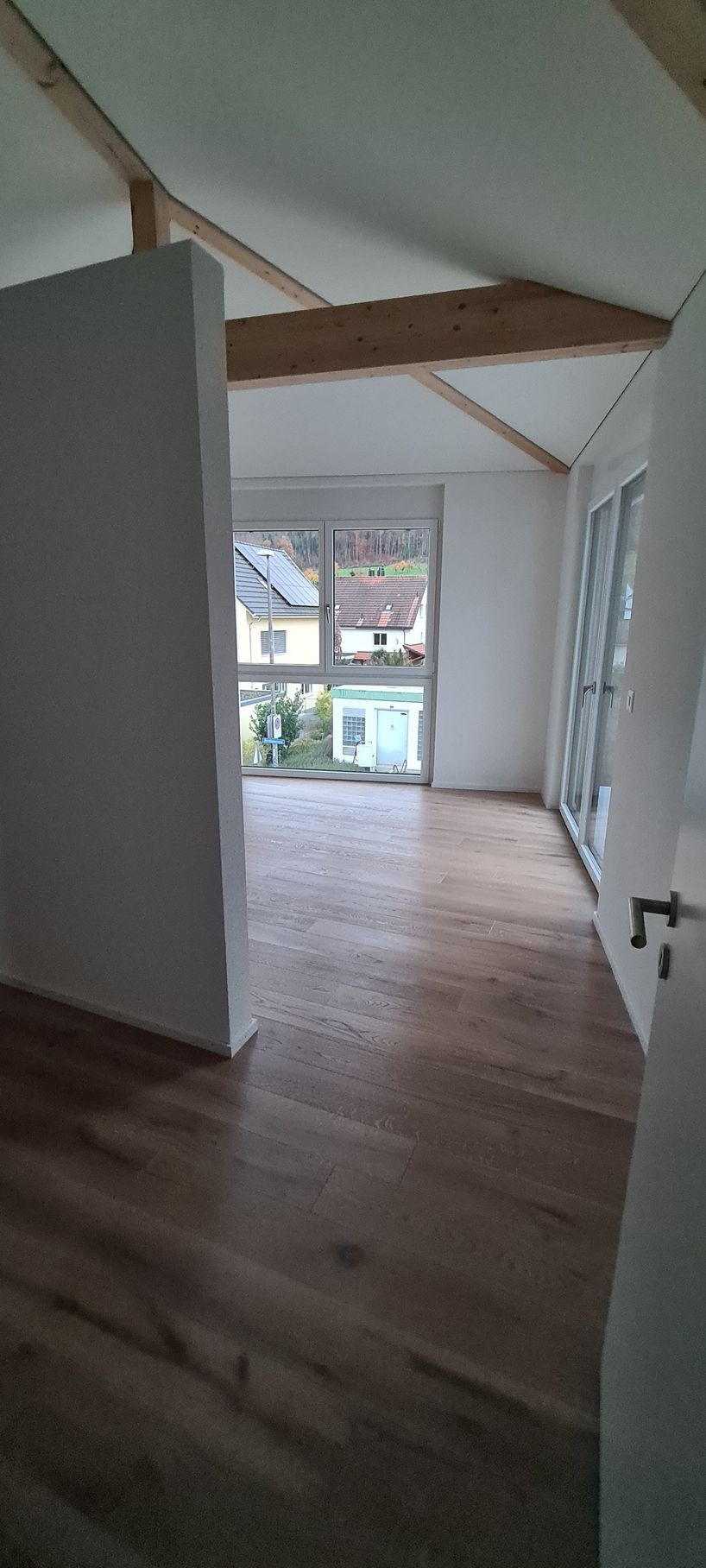 Innenansicht eines Raumes mit Holzboden, weißen Wänden, einem Fenster und einer Tür.