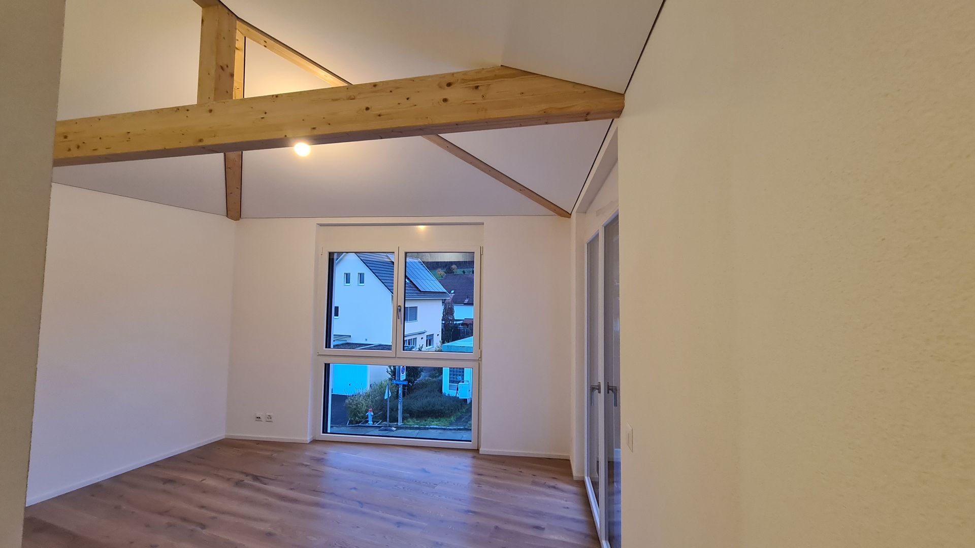 Leerer Raum mit Dachschräge, freiliegenden Holzbalken, Fenster mit Aussicht und Holzboden.