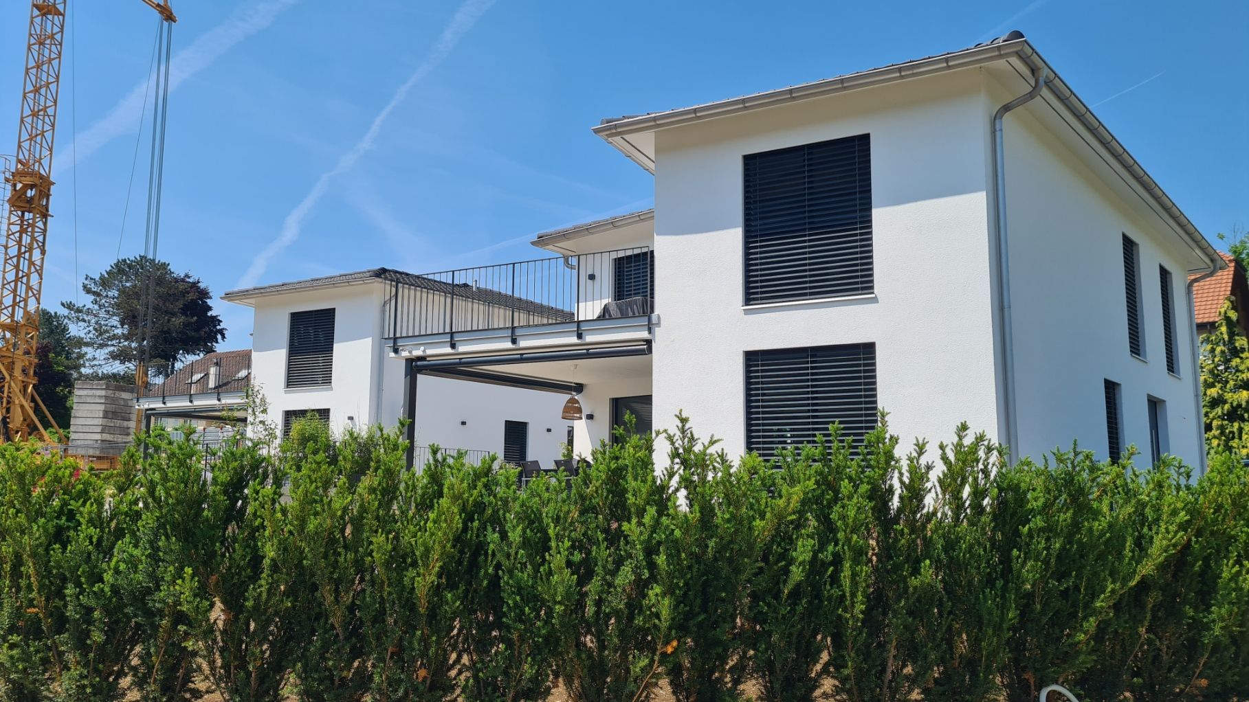 Weiße zweistöckige Häuser mit schwarzen Fensterläden hinter einer grünen Hecke unter blauem Himmel, mit einem Kran.