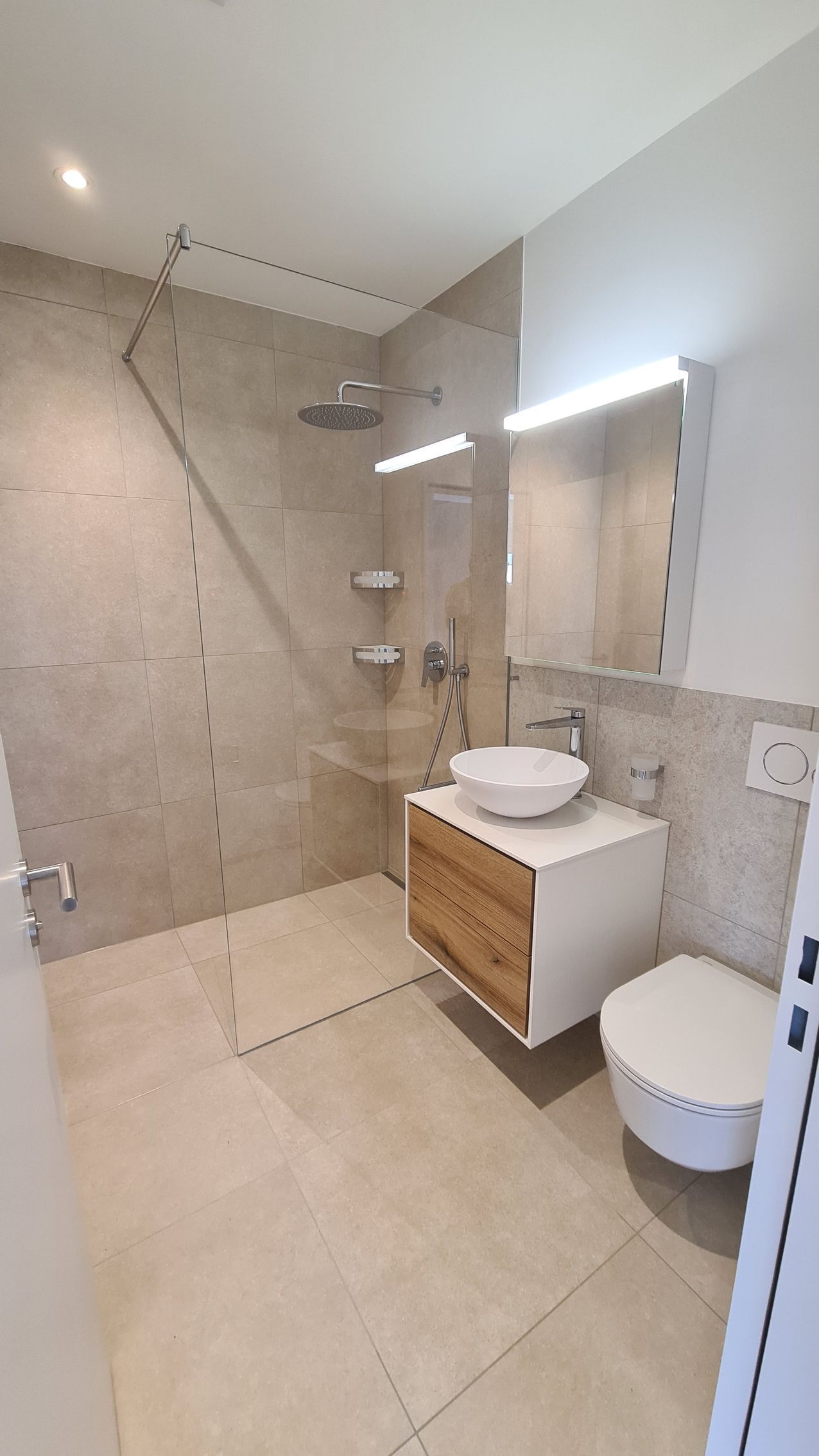 Modernes Badezimmer mit ebenerdiger Dusche, weißem Waschtisch und Toilette. Neutrale Fliesen und Holzakzente.