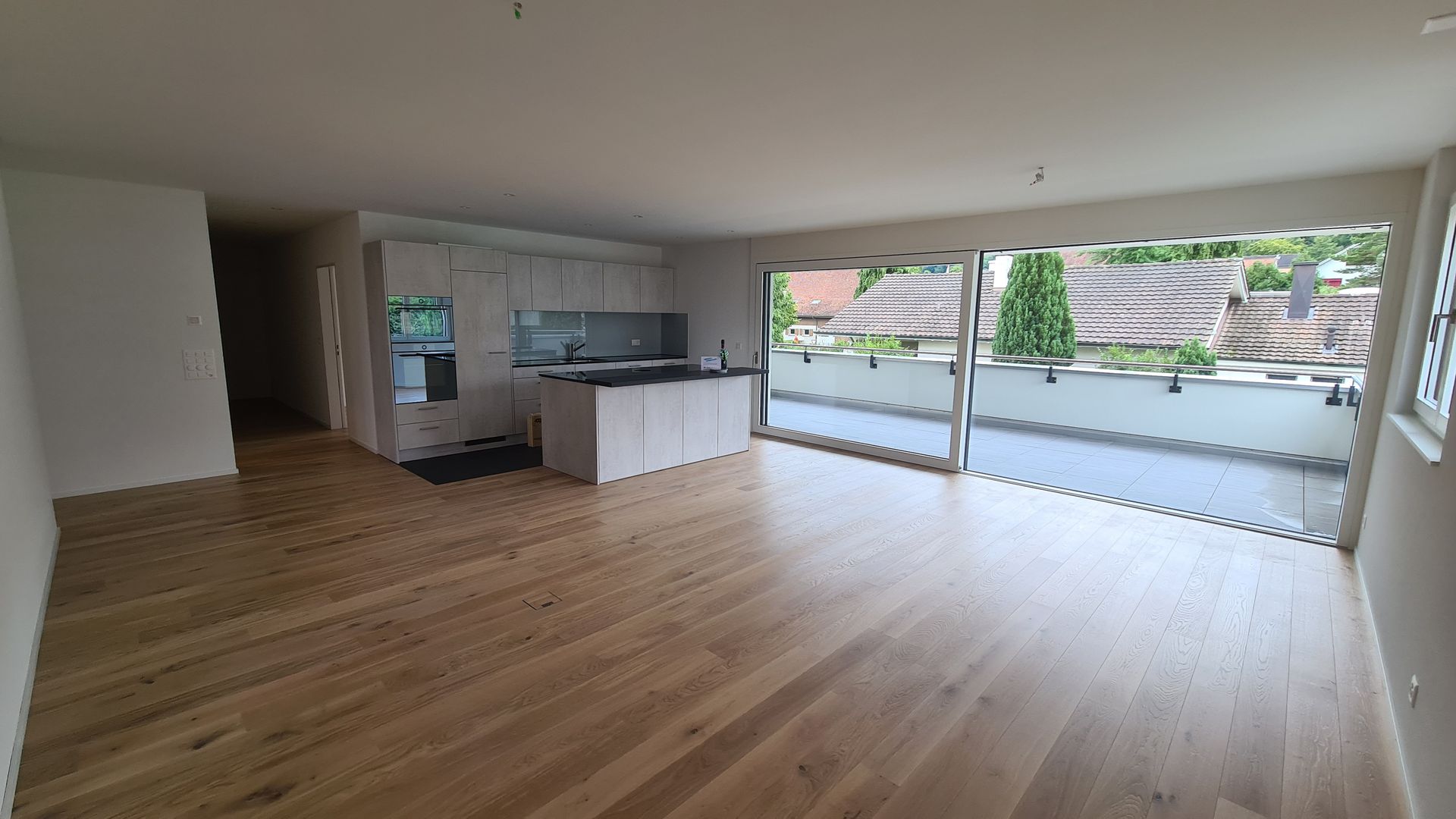 Leerer, offener Wohnbereich mit hellem Holzboden, weißen Schränken und Glasschiebetüren zu einer Terrasse.