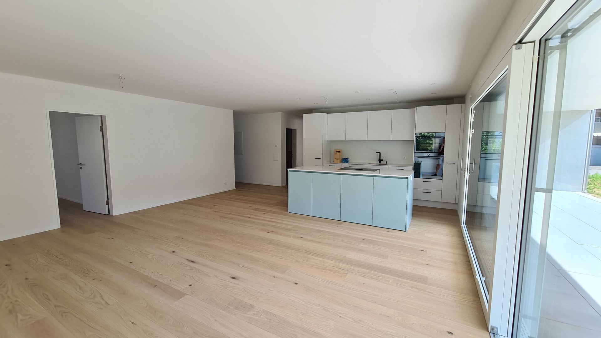 Offene Küche mit hellen Holzböden, weißen Wänden und hellblauer Kochinsel; große Glastüren zum Balkon.