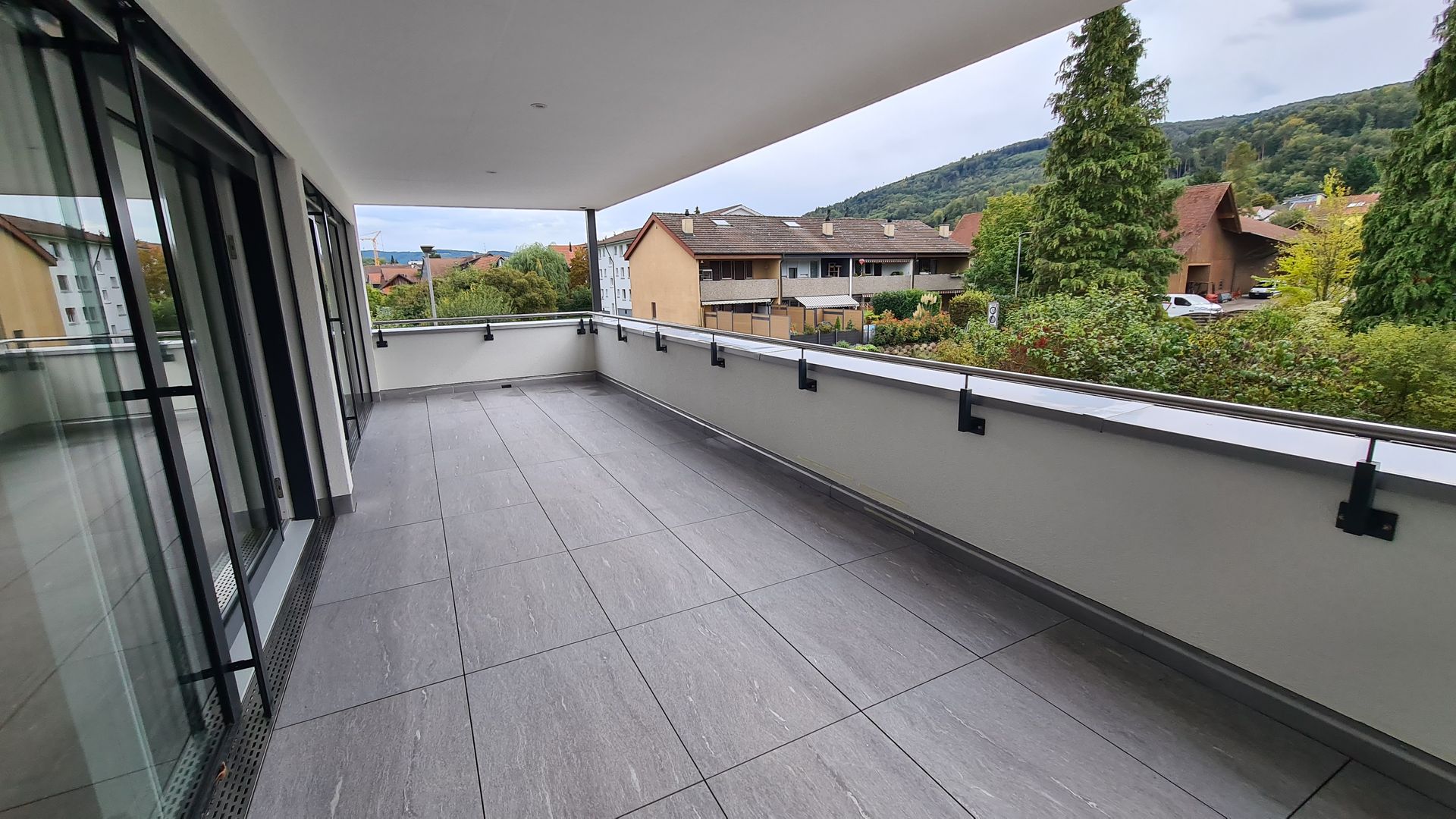 Balkon mit grauem Fliesenboden und weißem Geländer mit Blick auf Gebäude und Bäume.