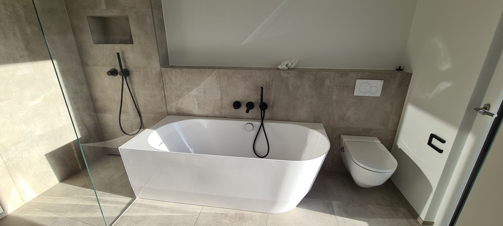Ein modernes Badezimmer mit weißer Badewanne, Toilette und Dusche. Die Wände sind in einer neutralen Farbe gefliest.