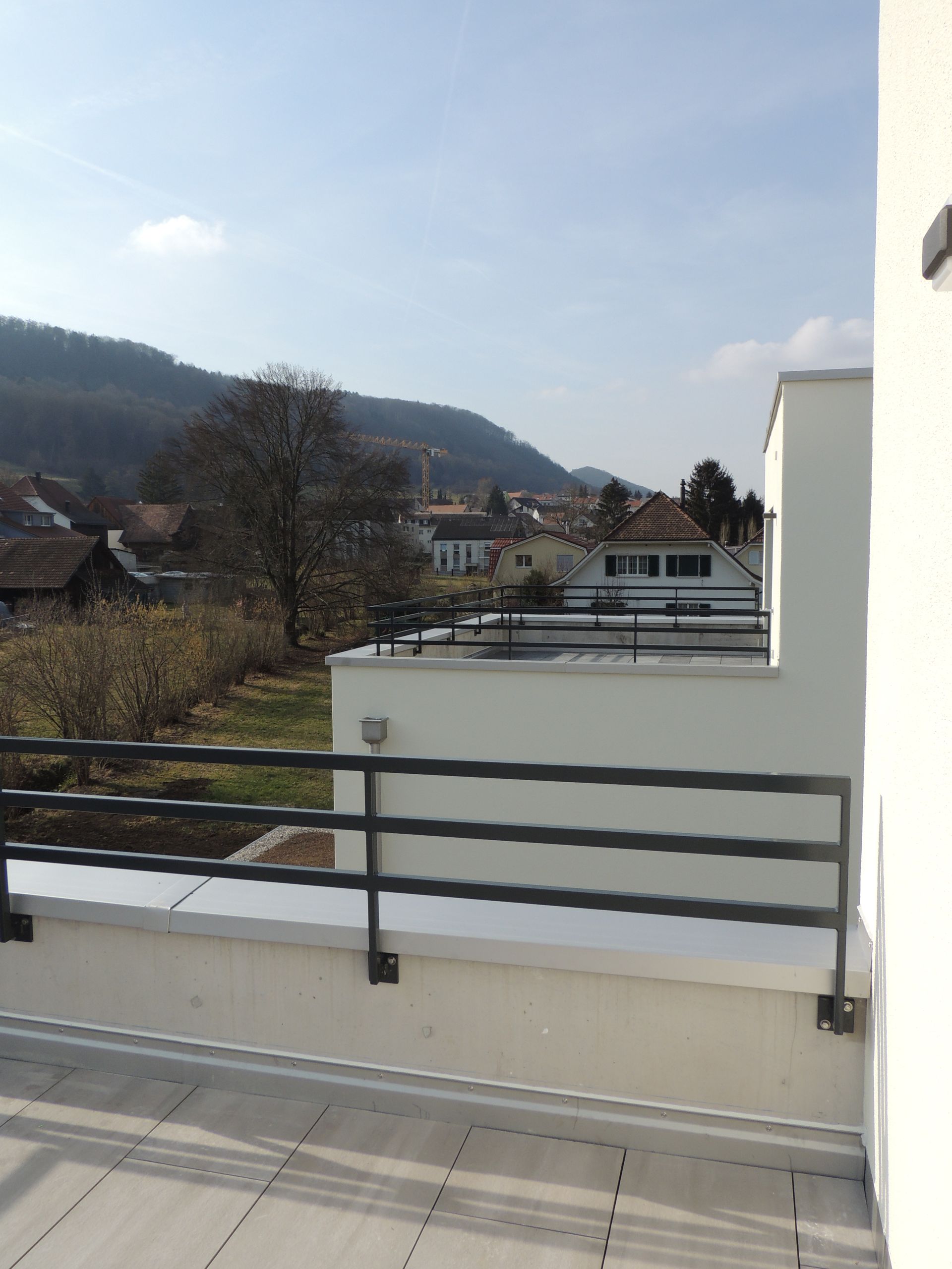 Blick von einer Dachterrasse mit Blick auf ein Dorf und Berge, mit weißen Wänden und schwarzem Geländer.