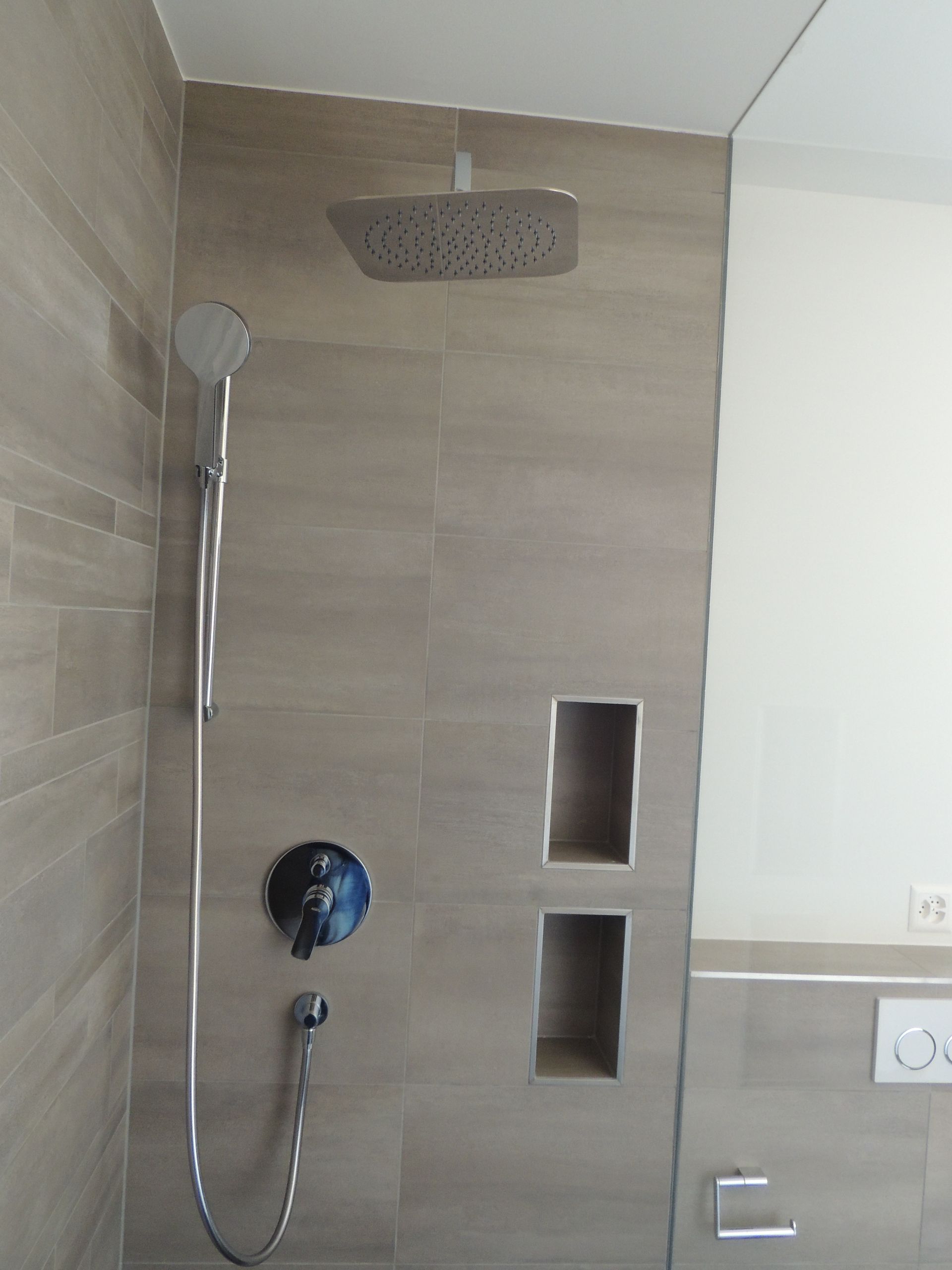 Dusche mit gefliester Wand, Kopfbrause, Handbrause, vertieften Ablagen und blauem Wasserhahn.