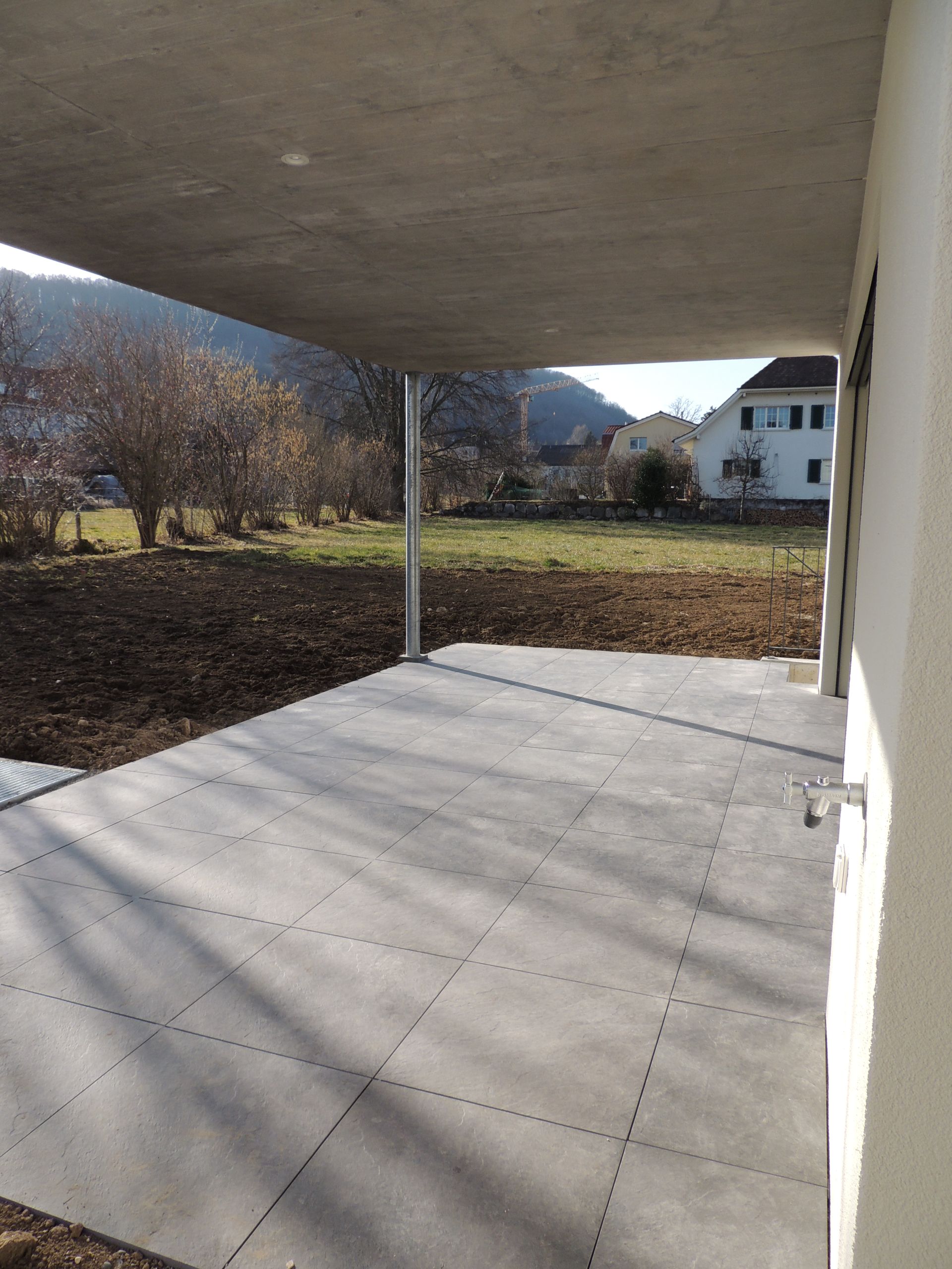Überdachte Terrasse mit grau gefliestem Boden, Metallstützpfosten und Blick auf einen Hof und entfernte Gebäude.