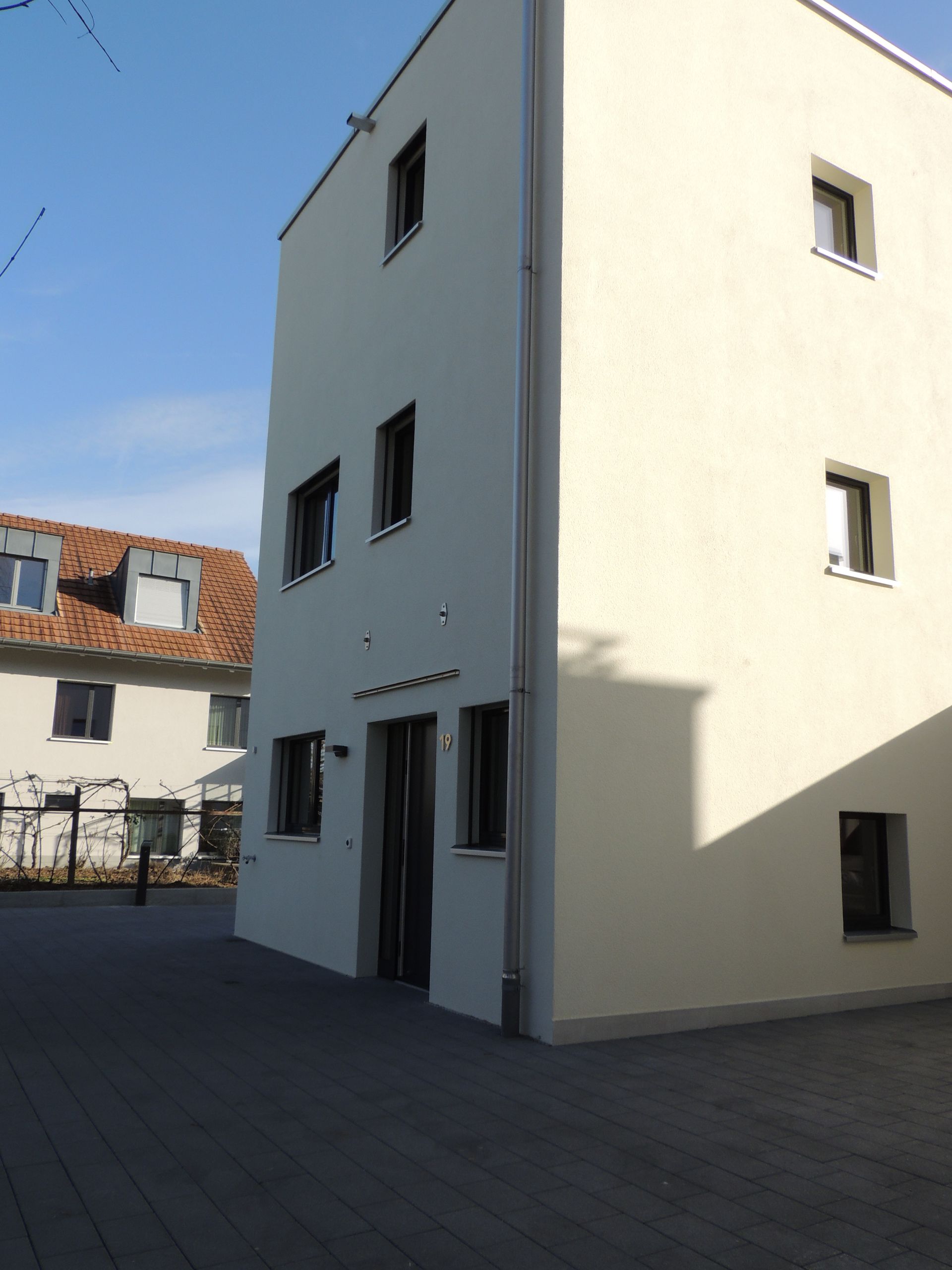 Beiges dreistöckiges Gebäude mit dunklen Fenstern, neben einem Backsteingebäude unter einem strahlend blauen Himmel.