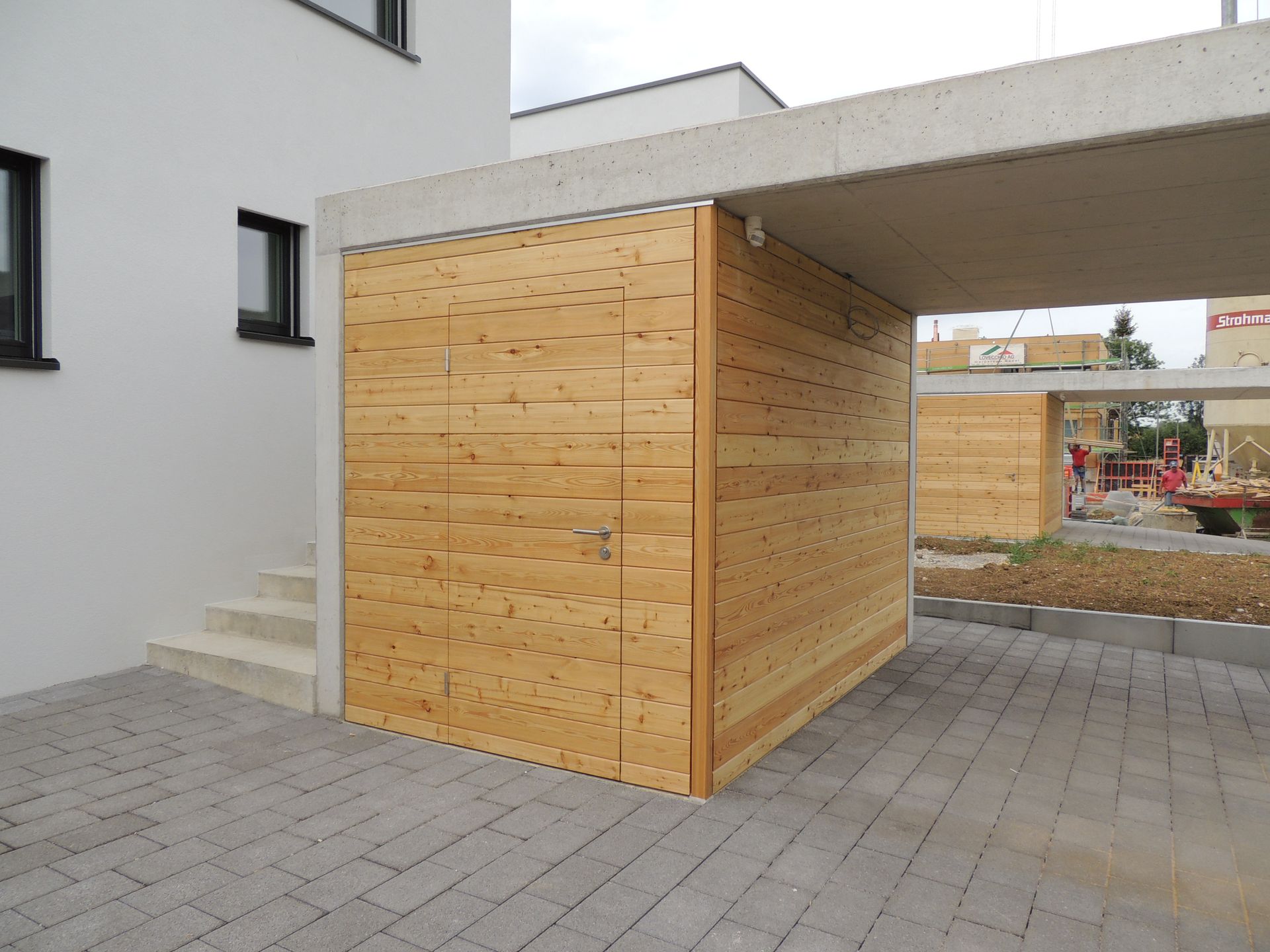Holzlagereinheit unter einem Betonüberhang neben einem Gebäude mit Treppe; Ziegelpflaster.