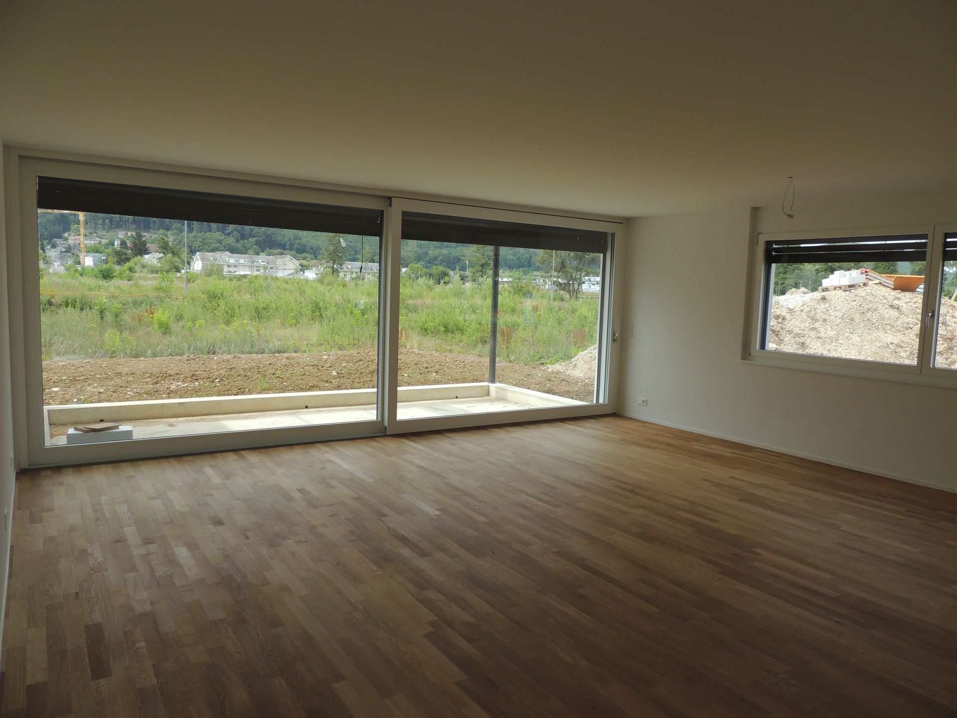 Leerer Raum mit Holzböden und großen Fenstern mit Blick auf einen Außenbereich.