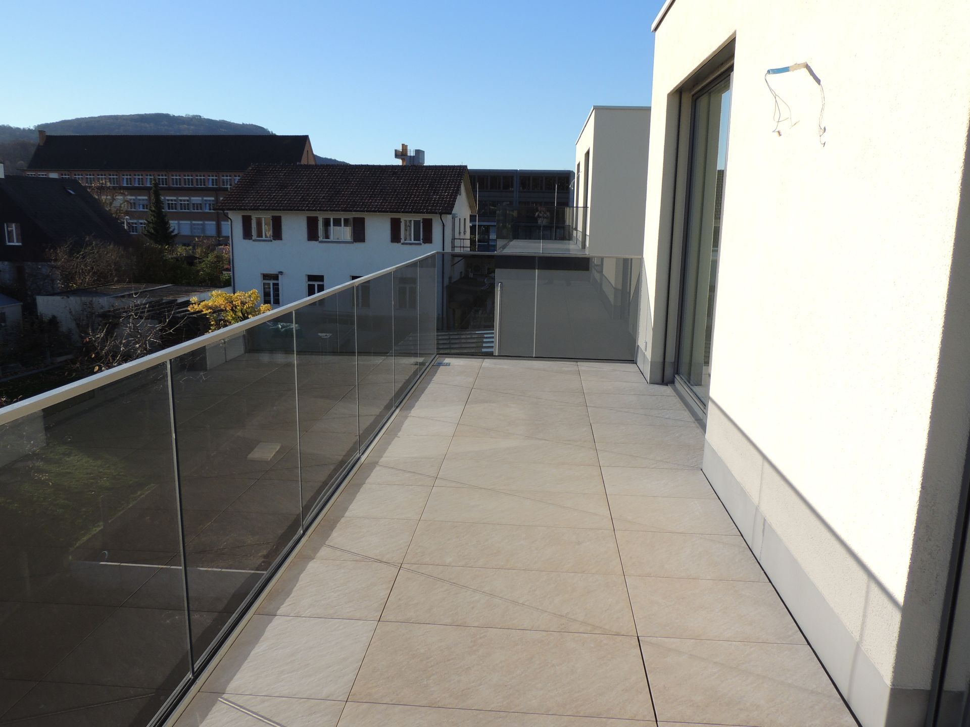 Balkon mit Glasgeländer, hellbraunem Fliesenboden und Blick auf Gebäude und Berge im Hintergrund.
