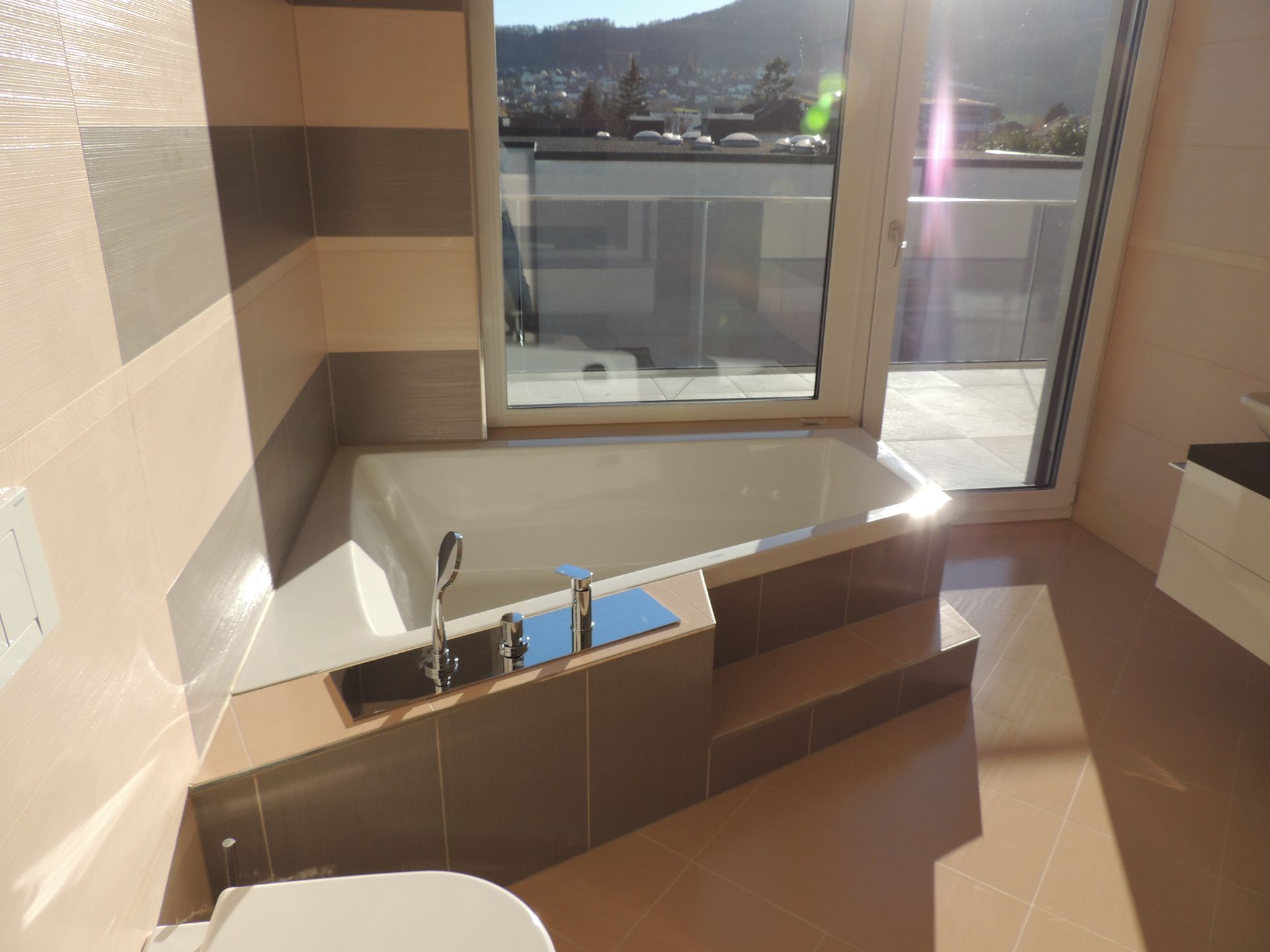 Badezimmer mit eingebauter Badewanne am Fenster. Hellbraune und graue Fliesen, Sonnenlicht.