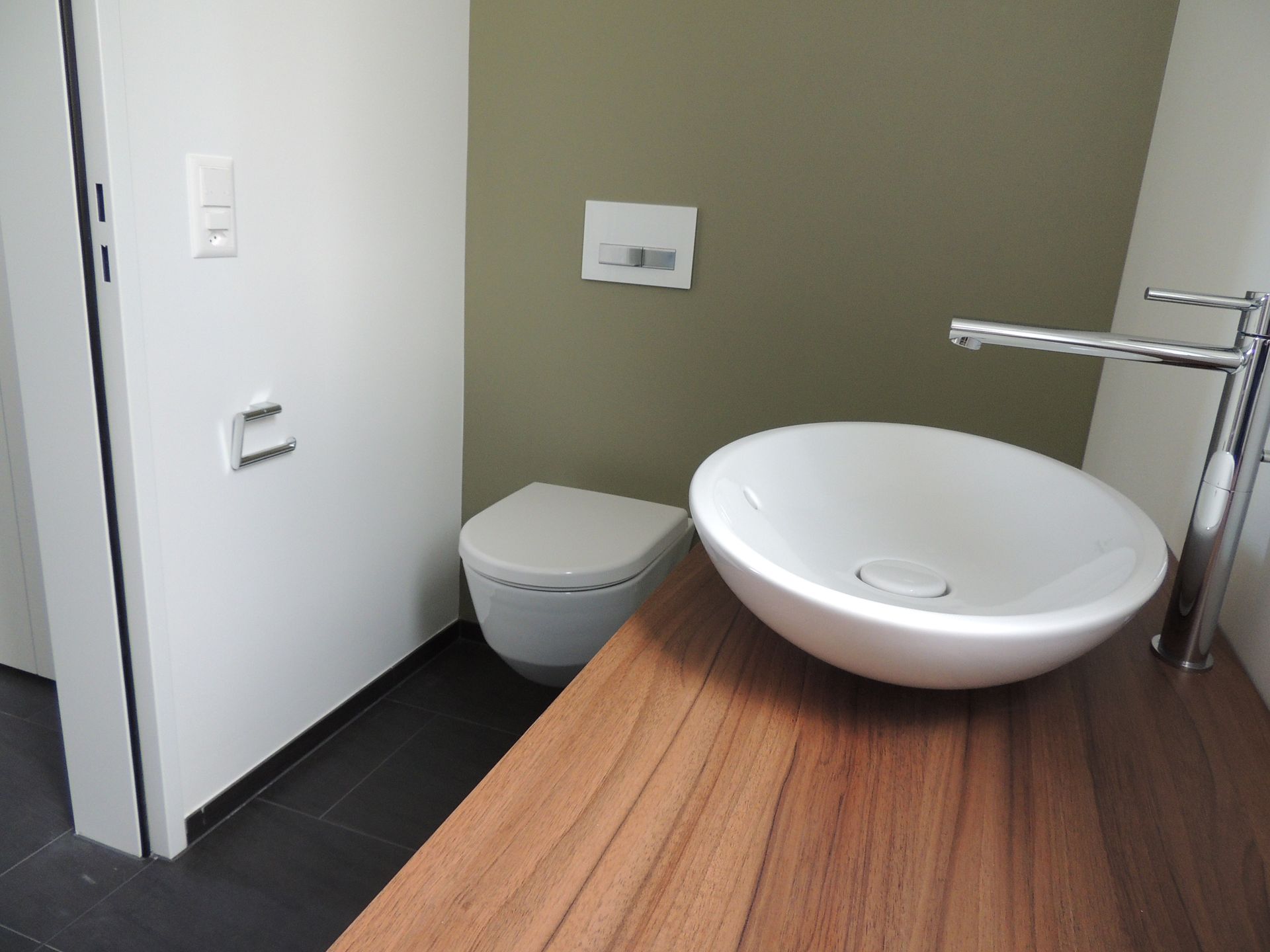 Badezimmer mit einem weißen ovalen Waschbecken auf einer Holzablage, einer Toilette und einer olivgrünen Wand.
