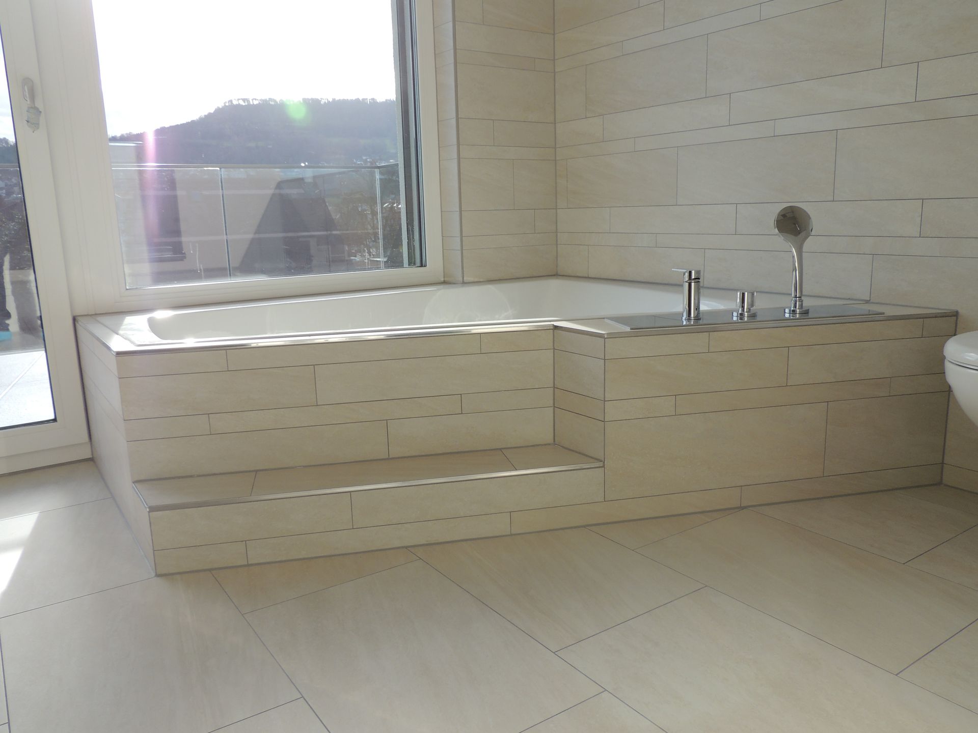 Weiß gefliestes Badezimmer mit eingebauter Badewanne, Stufen und großem Fenster mit Blick auf einen Hügel.