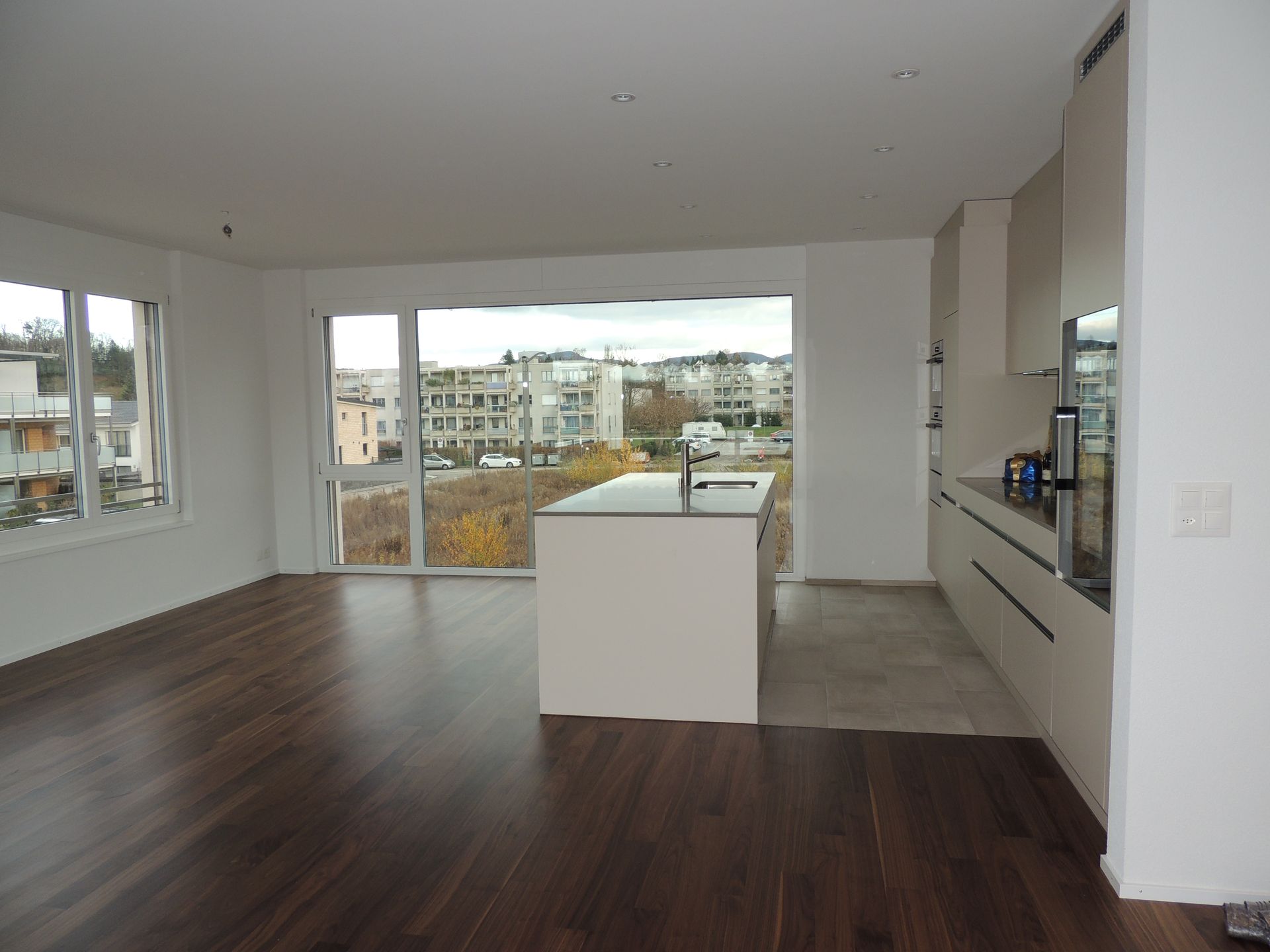 Moderne offene Küche/Wohnbereich mit dunklen Holzböden, großem Fenster mit Blick auf Gebäude, weißen Schränken, Kochinsel.