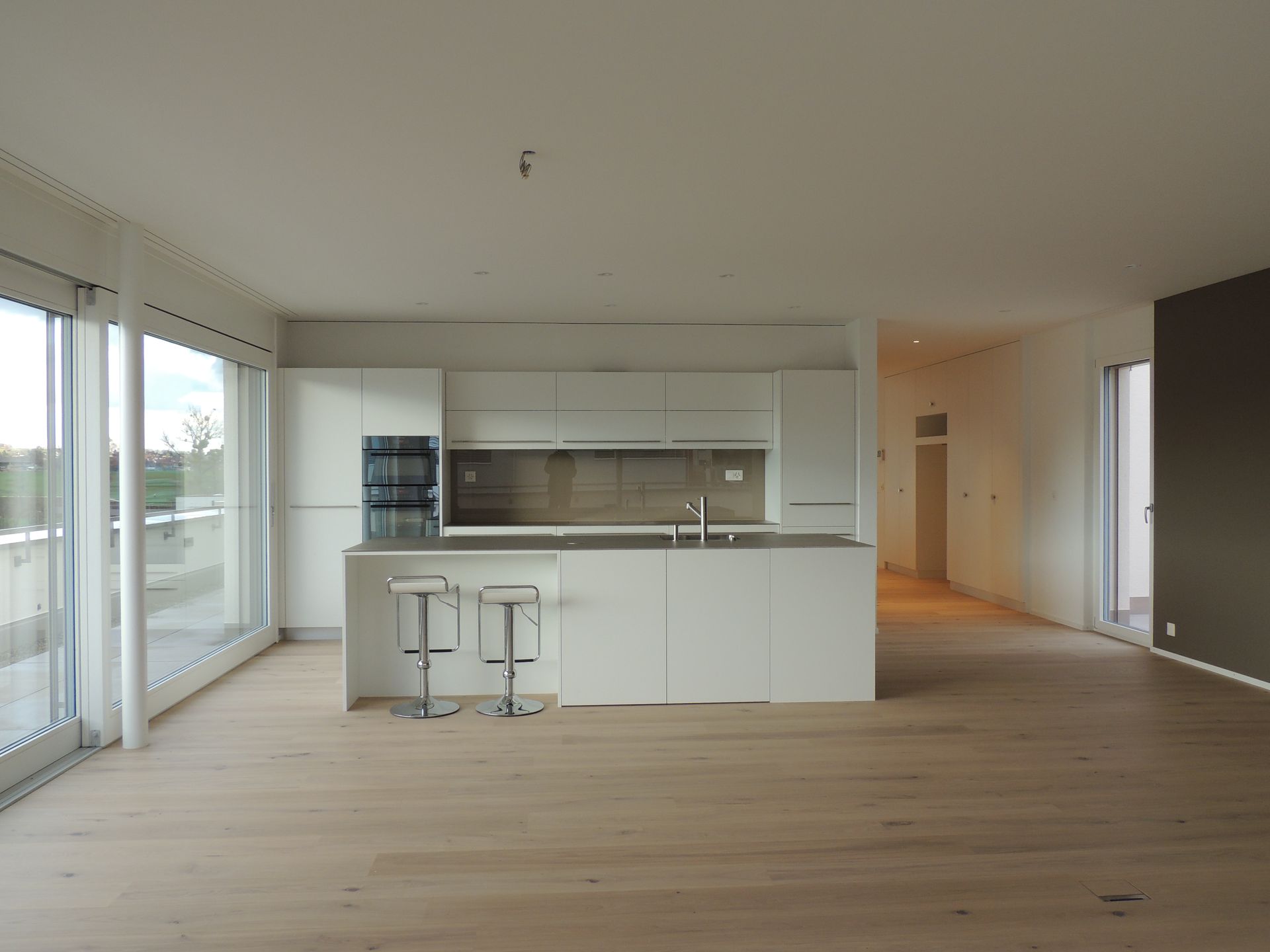 Moderne offene Küche mit weißen Schränken, Geräten aus Edelstahl und einer grauen Kochinsel. Heller Holzboden, Glasschiebetüren.