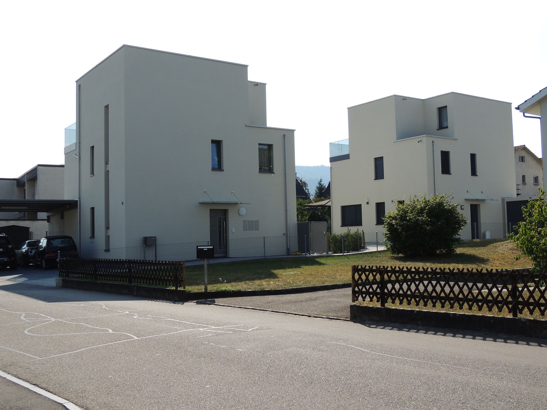 Zwei moderne, helle, quadratische Häuser mit kleinen Fenstern und einem grasbewachsenen Hof, getrennt durch einen dunklen Zaun und eine Straße.