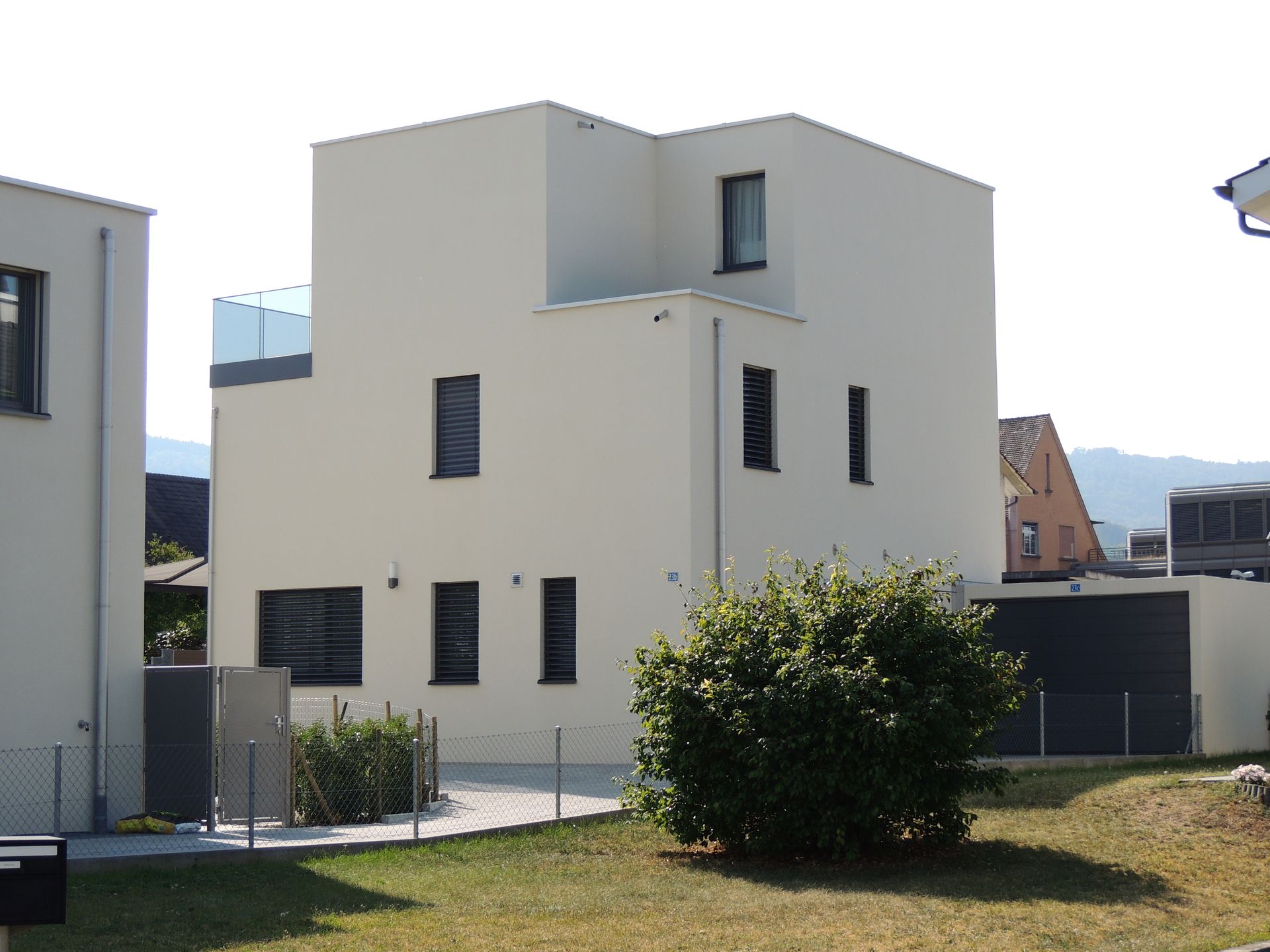 Modernes weißes Haus mit dunklen Fenstern, kleinem Balkon und gepflegtem Rasen unter einem strahlenden Himmel.