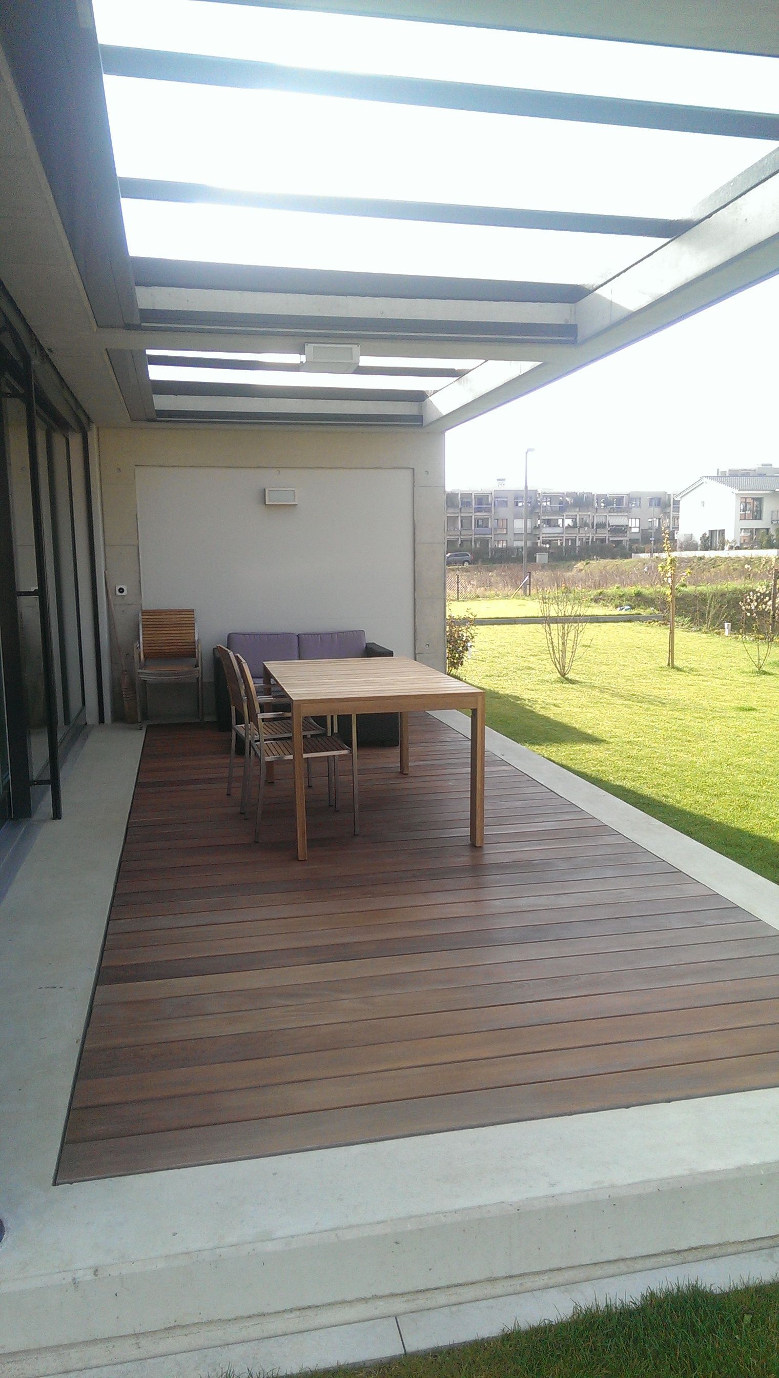 Außenterrasse mit Holzdeck, Tisch, Stühlen und grünem Rasen. Sonnenlicht scheint durch die Überkopfkonstruktion.
