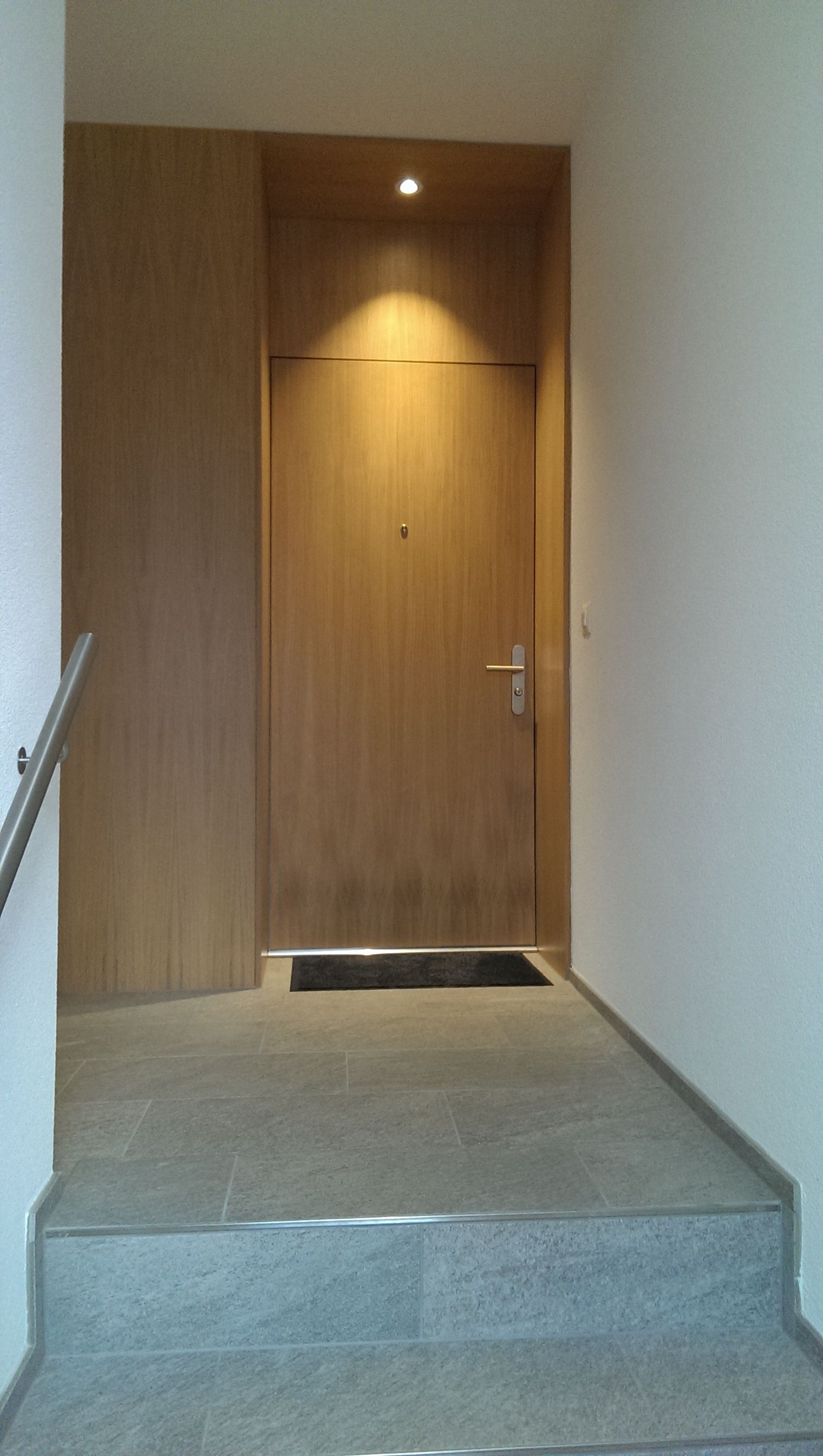 Eine Holztür in einem hellen Eingangsbereich. Stufen führen hinauf. Helle Wände und Böden.