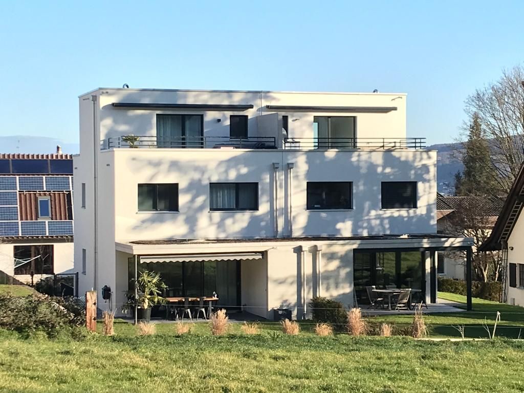 Modernes dreistöckiges weißes Gebäude mit Balkonen und großen Fenstern, inmitten einer Rasenfläche mit Bäumen.