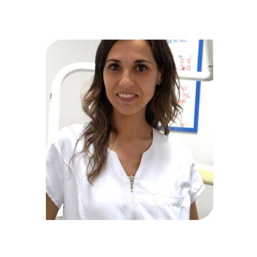 Mujer con uniforme blanco de pie en una consulta dental, sonriendo a la cámara.