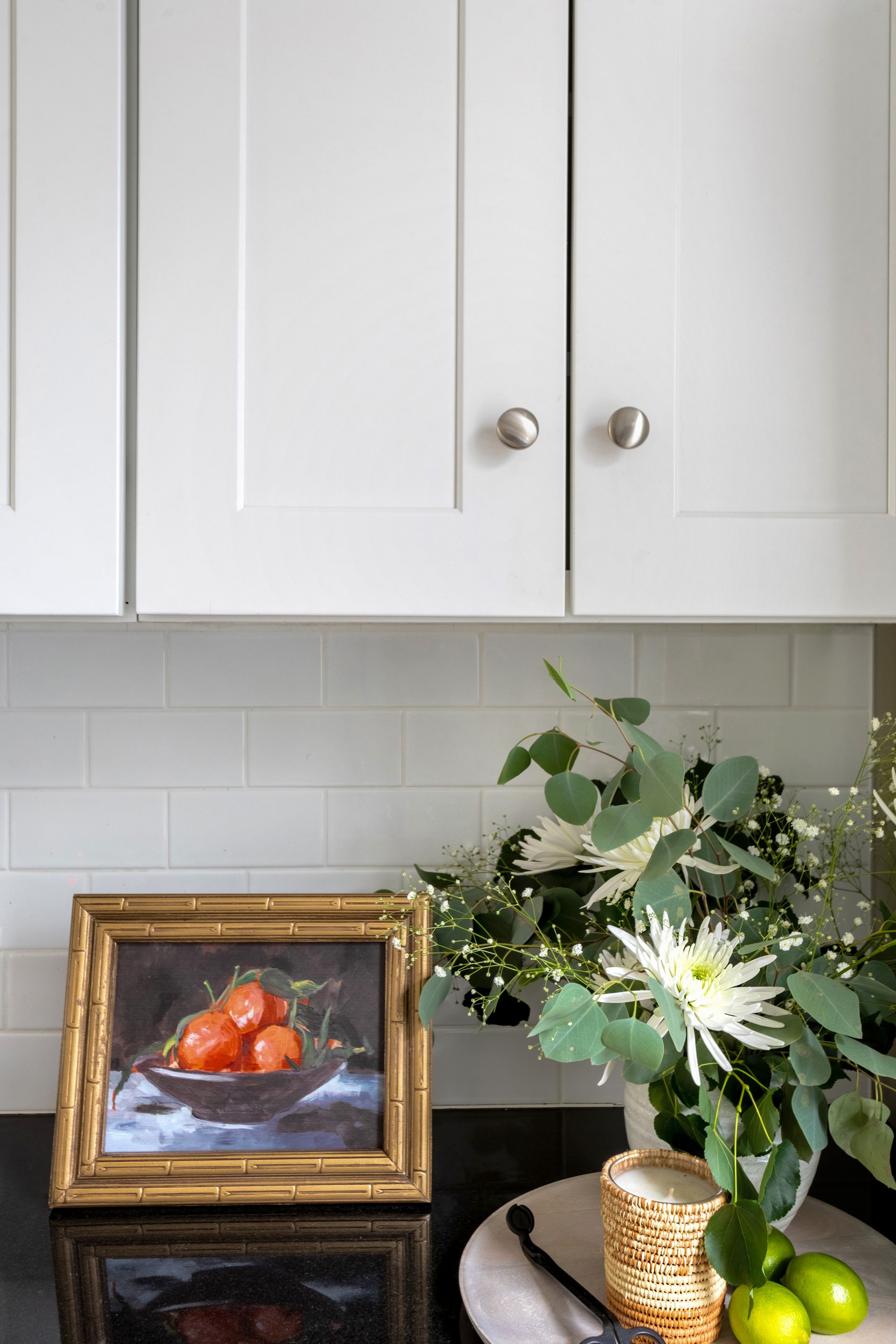 A painting of tomatoes in a bowl is on a kitchen counter next to a vase of flowers.