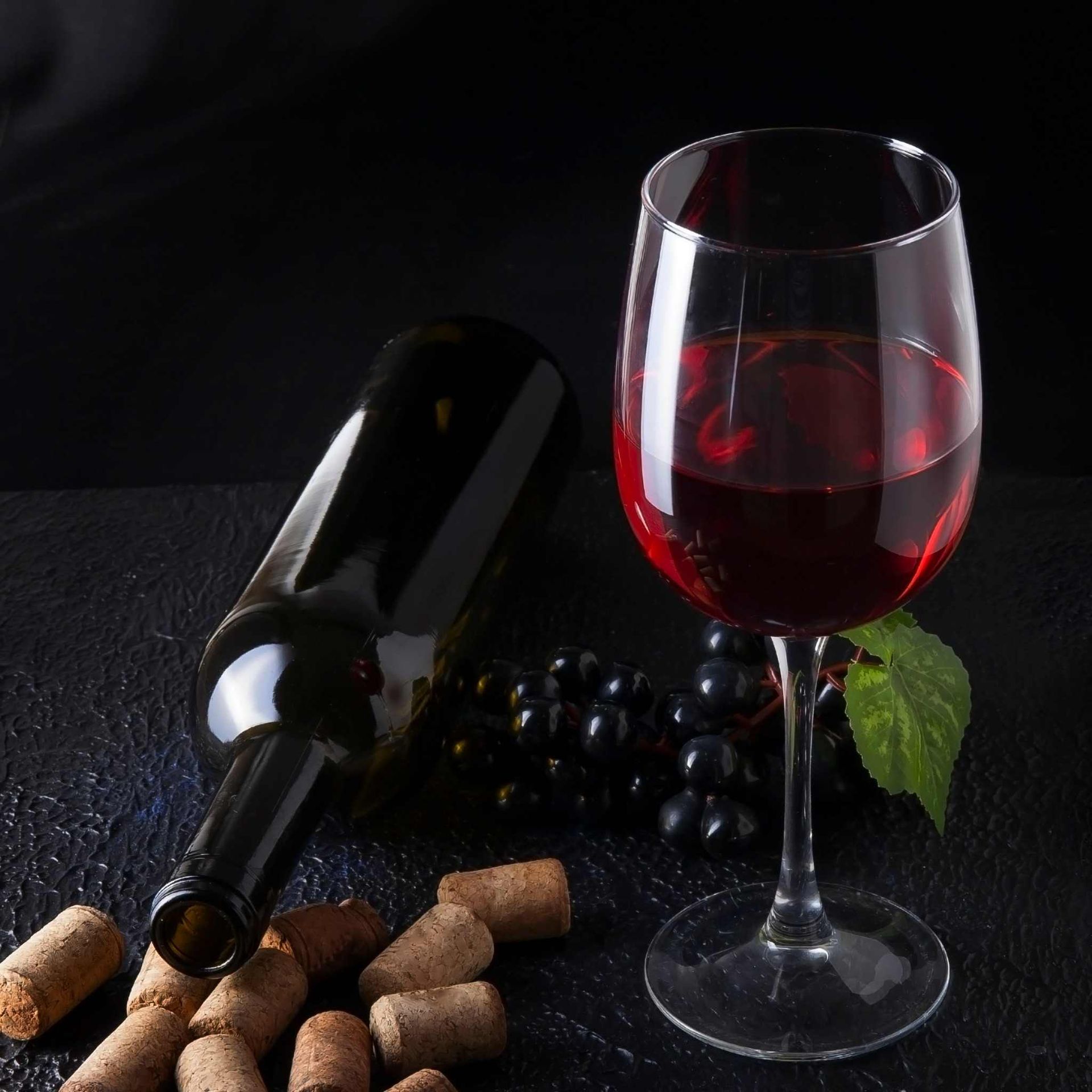 Una copa de vino tinto y una botella sobre una superficie oscura, con uvas y corchos.