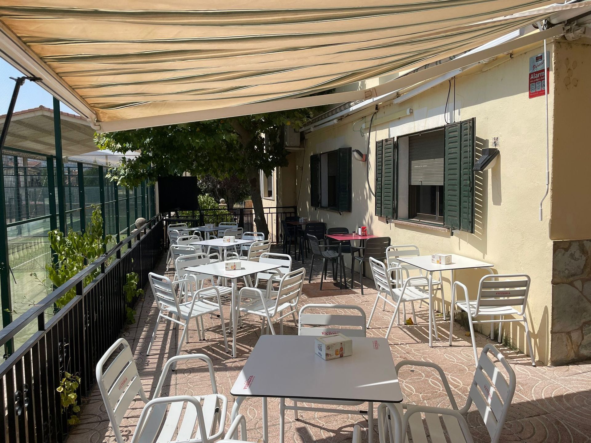 Cafetería al aire libre con mesas y sillas blancas bajo un toldo beige.
