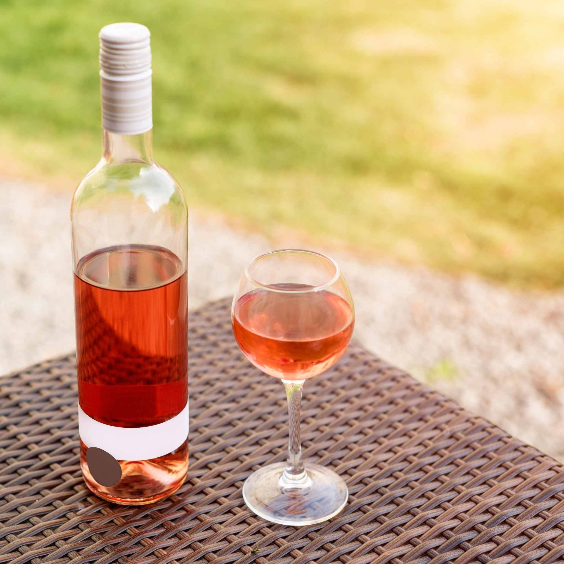 Botella y copa de vino rosado sobre una mesa tejida al aire libre, con un fondo verde borroso.