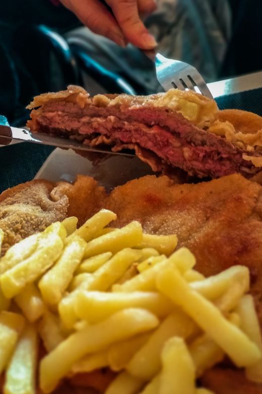 Primer plano de un plato con una persona sosteniendo un tenedor en una rebanada de carne frita. Incluye papas fritas y otros alimentos fritos.