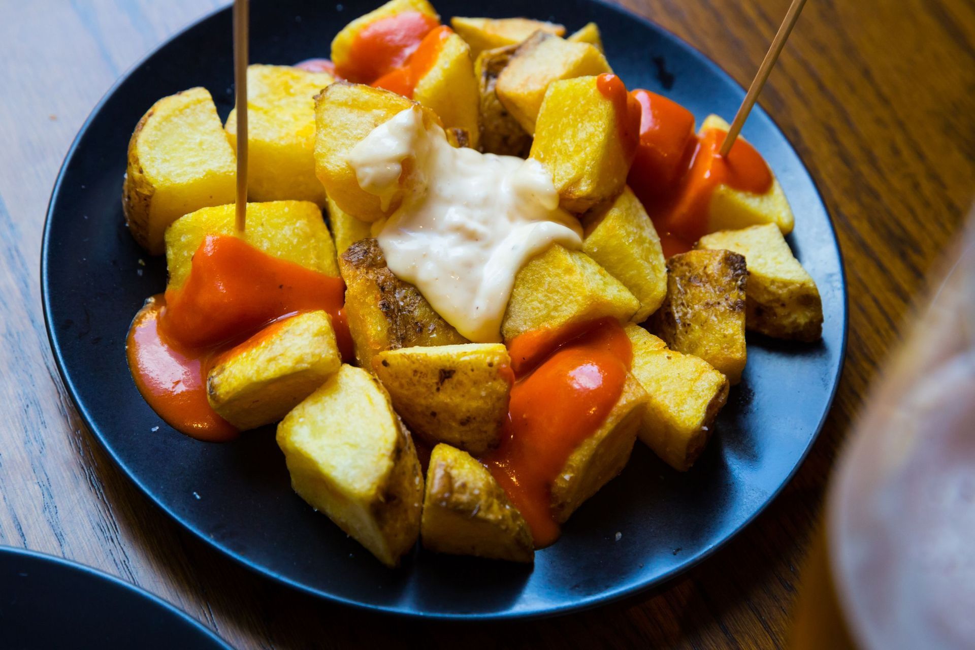 Patatas bravas: cubos de patata fritos con salsas roja y blanca en un plato oscuro, sujetos con palillos.