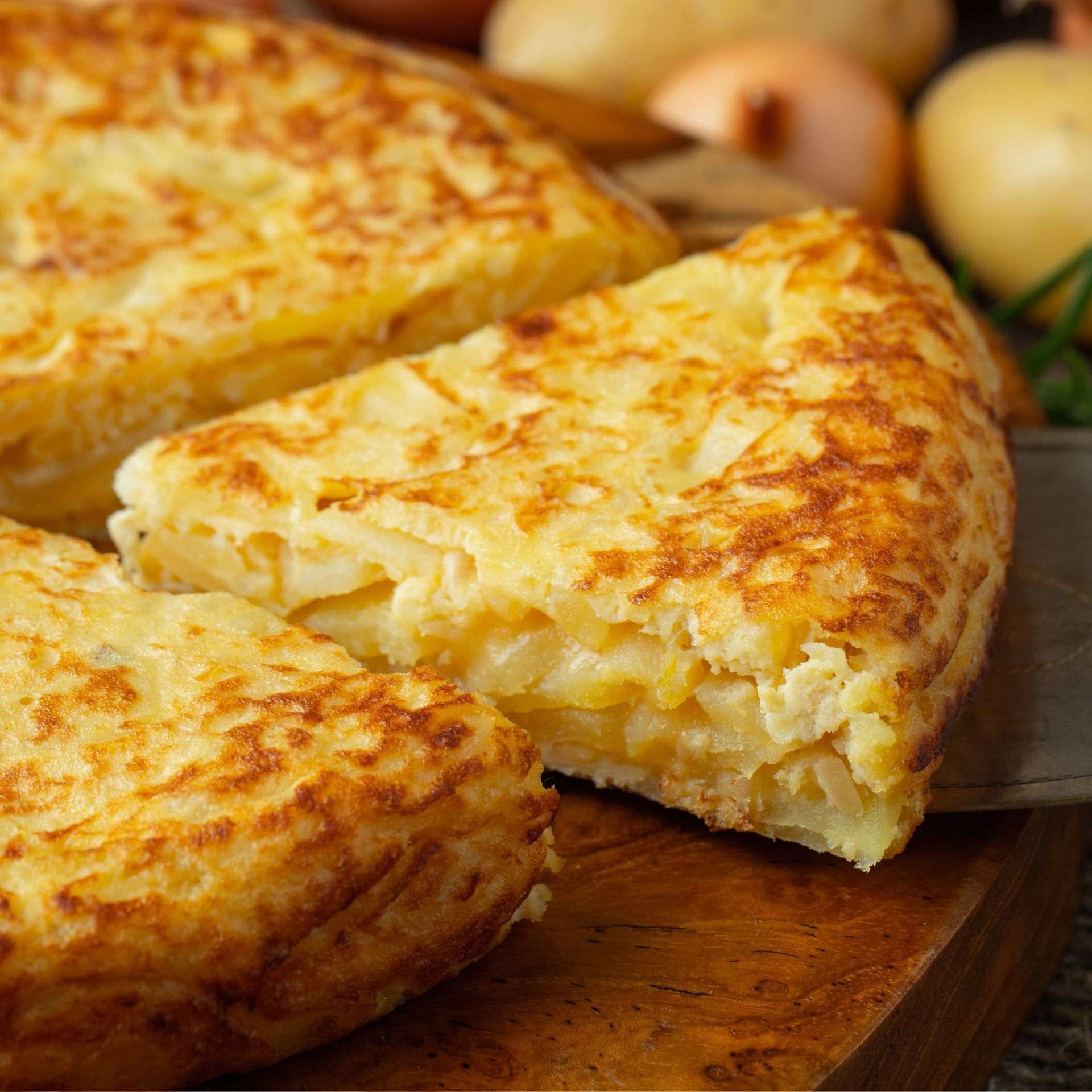 Tortilla española, dorada con la rodaja cortada, sobre una tabla de madera, con patatas y cebollas al fondo.