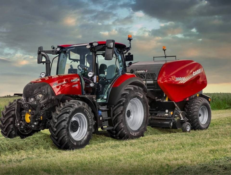 Ein roter Traktor zieht ein rotes landwirtschaftliches Gerät über ein grasbewachsenes Feld unter einem bewölkten Himmel