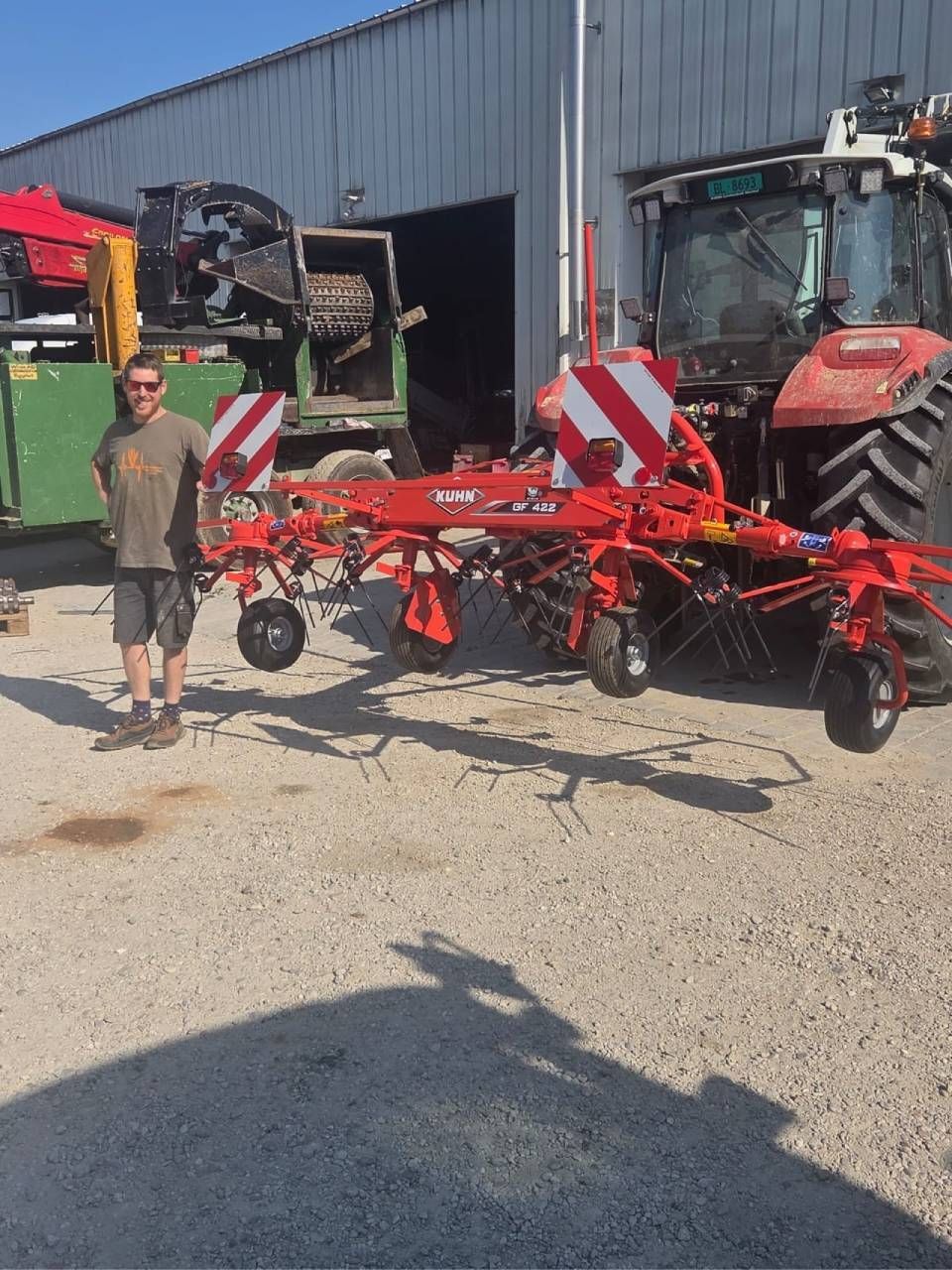 Eine Person steht neben einem roten landwirtschaftlichen Gerät vor einer Scheune und einem Traktor.