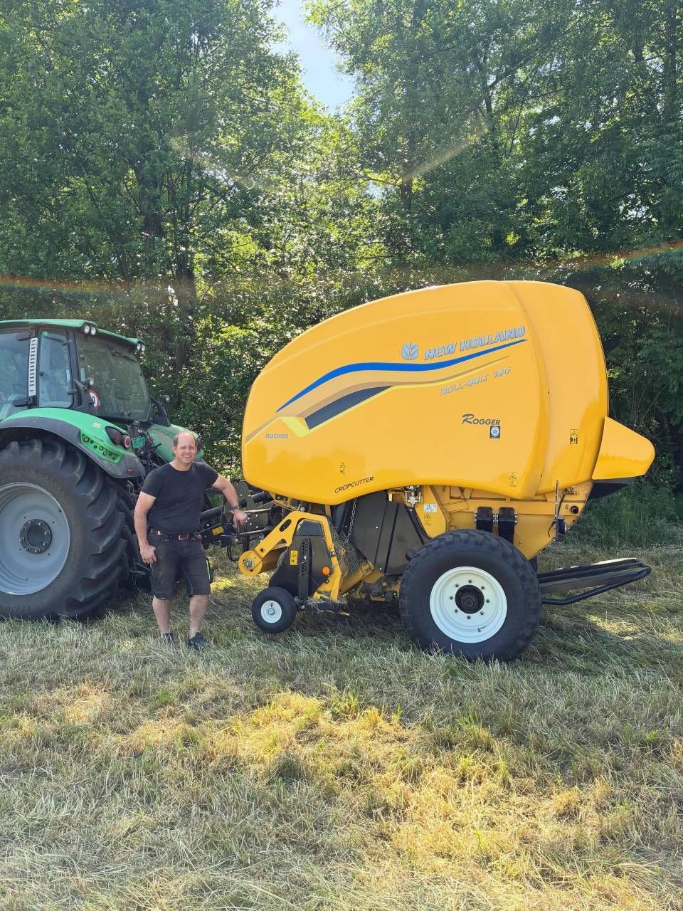 Eine gelbe Rundballenpresse steht neben einem Traktor auf einem grasbewachsenen Feld; eine Person steht in der Nähe unter Bäumen.