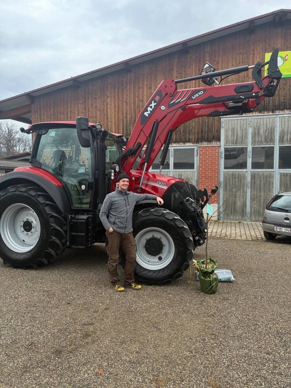 Eine Person steht neben einem roten Traktor mit Frontlader auf einem Bauernhof neben einer Scheune.