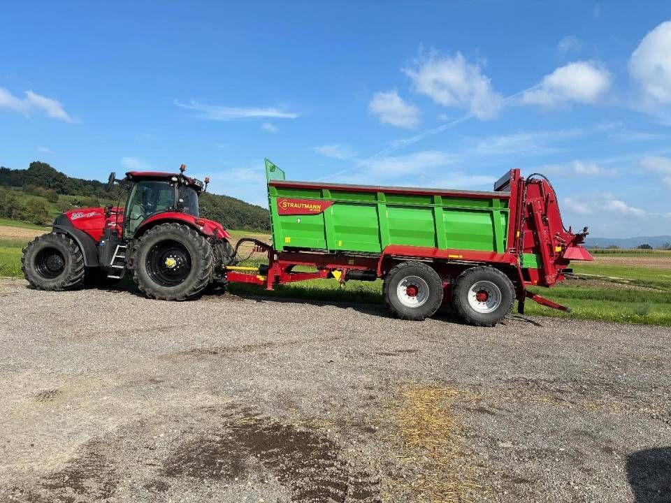 Ein roter Traktor zieht einen Gründüngungsstreuer auf einem Kiesfeld unter blauem Himmel