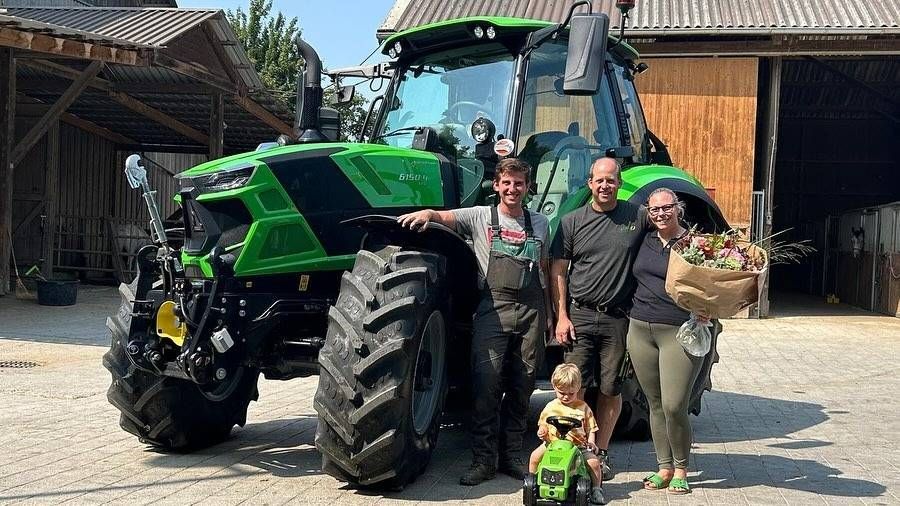 Menschen posieren neben einem grünen Traktor auf einem Bauernhof; ein Kind hält einen kleinen Spielzeugtraktor.