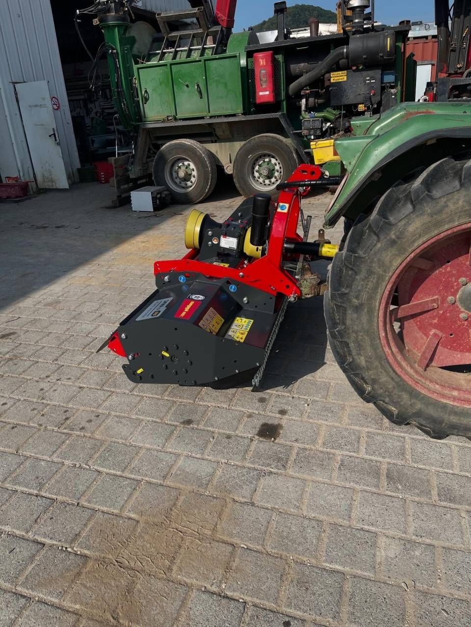 Ein roter, am Traktor montierter Mähaufsatz steht auf einer gepflasterten Fläche neben einem grossen grünen landwirtschaftlichen Fahrzeug.