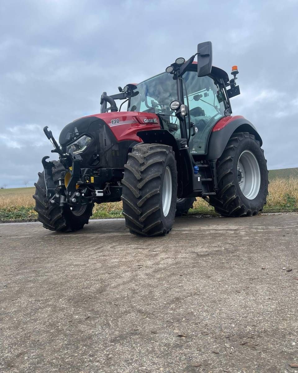 Ein roter Traktor mit Frontanbaugerät parkt auf einer Schotterstrasse in einer bewölkten Landschaft.