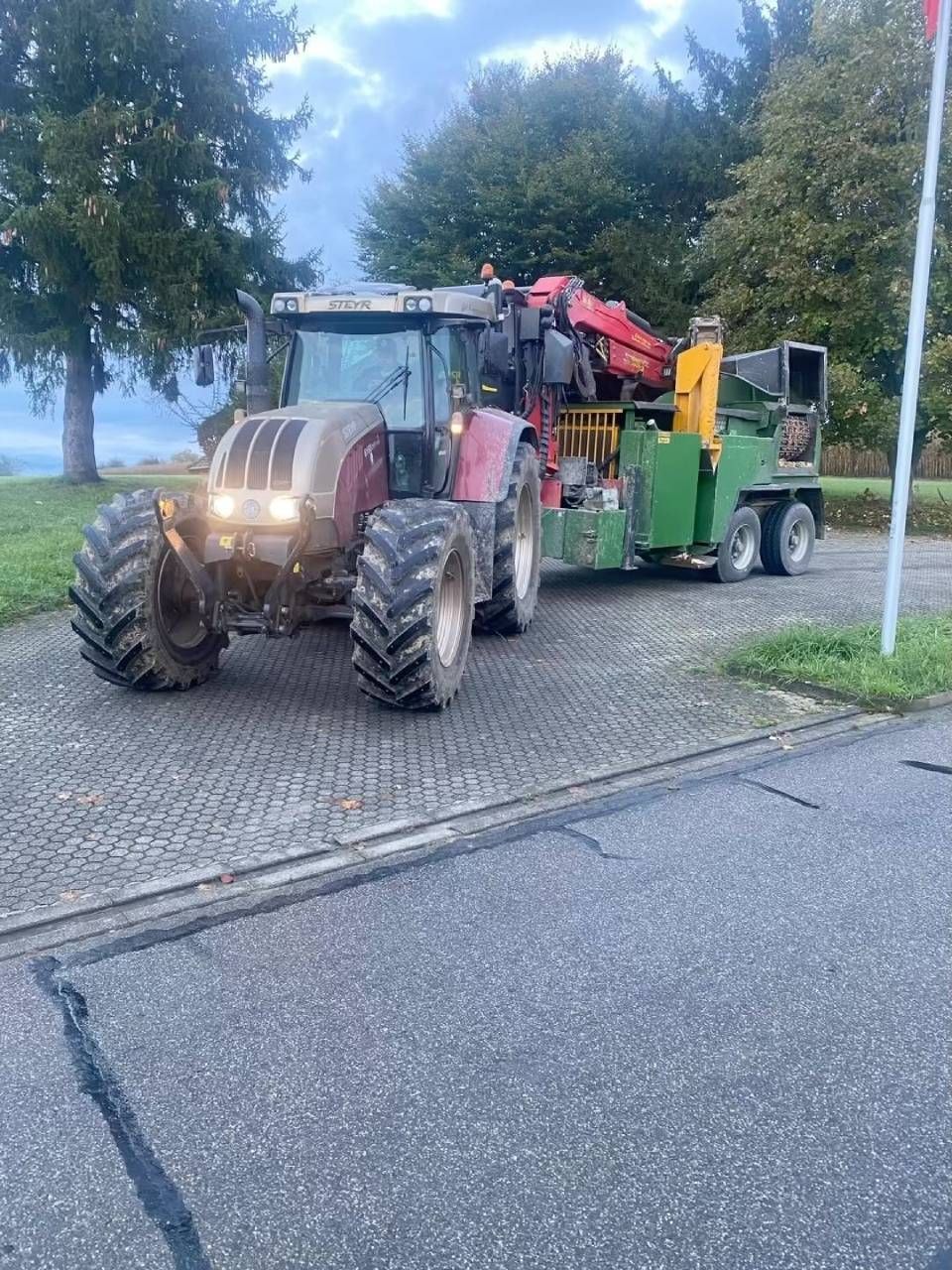 Ein roter Traktor zieht eine grüne Maschine auf einer Kopfsteinpflasterstrasse neben Bäumen und Wasser unter einem bewölkten Himmel