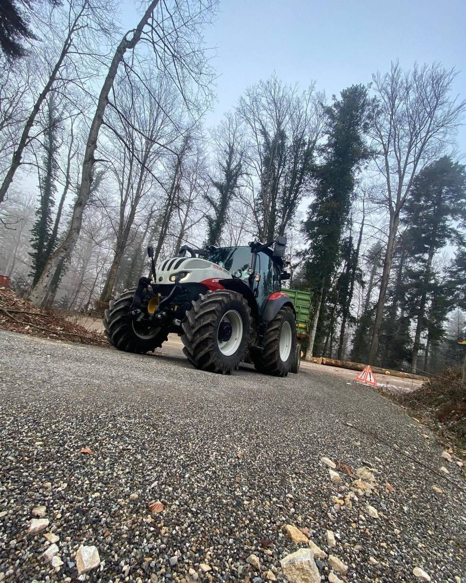 Blauer Traktor auf einer Schotterstrasse in einem Waldgebiet, aus niedriger Perspektive betrachtet.