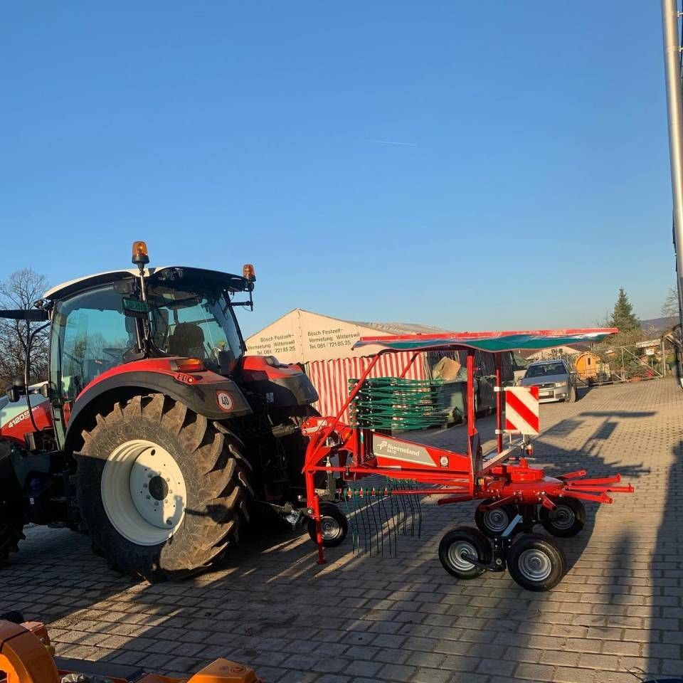Ein roter Traktor mit landwirtschaftlichen Geräten steht auf einem gepflasterten Parkplatz unter strahlend blauem Himmel.