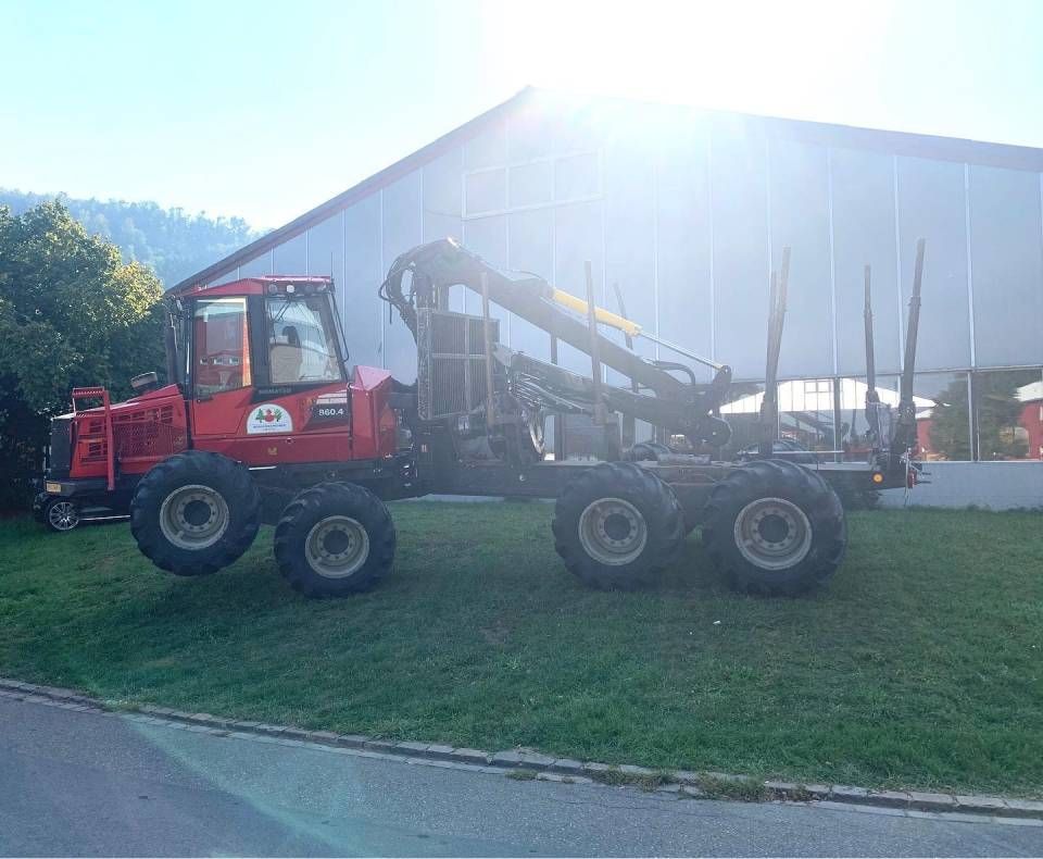 Ein roter Lkw zieht einen grossen Förderbandanhänger auf einer Wiese neben einem Lagerhaus im hellen Sonnenlicht.
