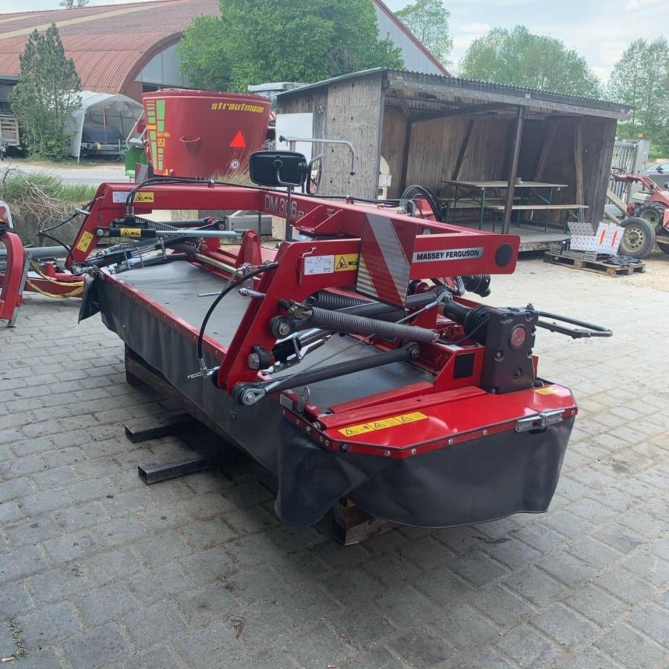 Ein roter Mähaufsatz für landwirtschaftliche Geräte steht auf einer gepflasterten Auffahrt; im Hintergrund ist ein roter Maschinenanhänger zu sehen.
