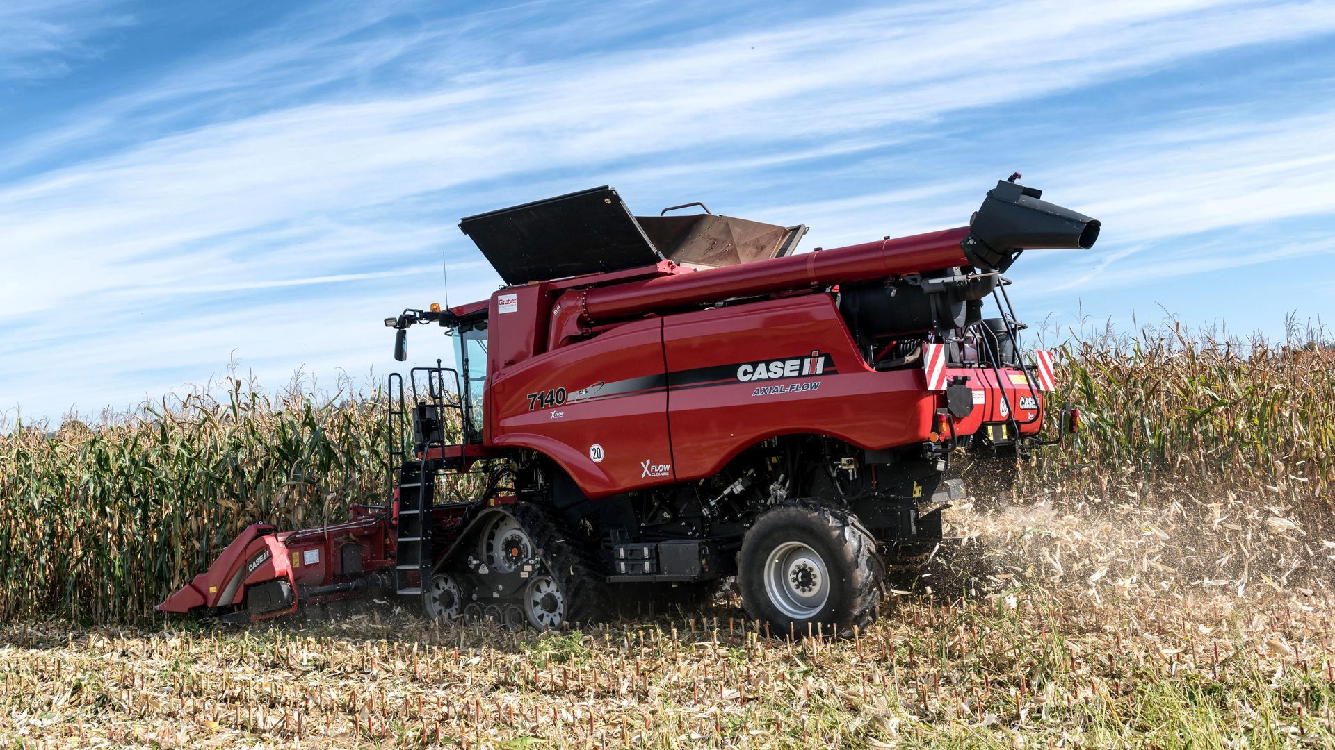 Eine Person steht neben einem roten landwirtschaftlichen Gerät vor einer Scheune und einem Traktor.